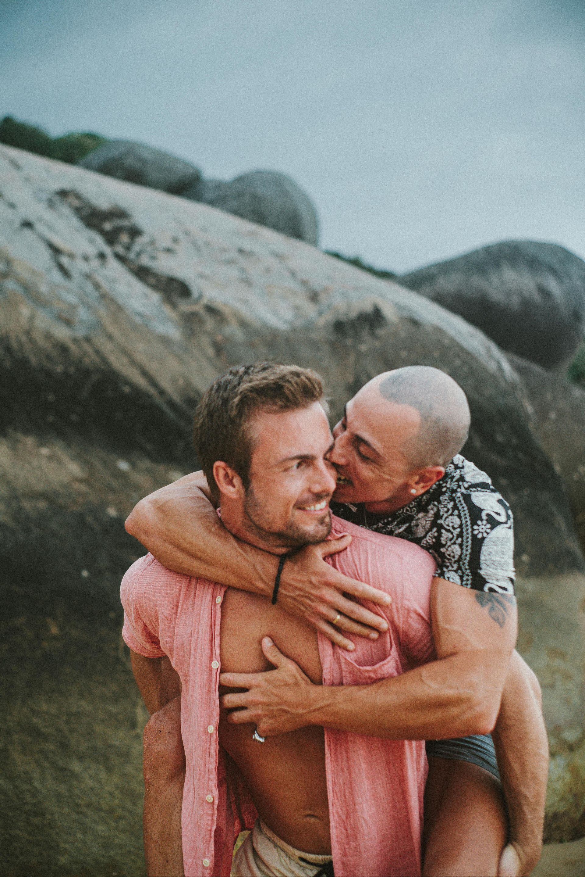 A person gives another person a piggyback ride on a rocky beach, smiling as they embrace.