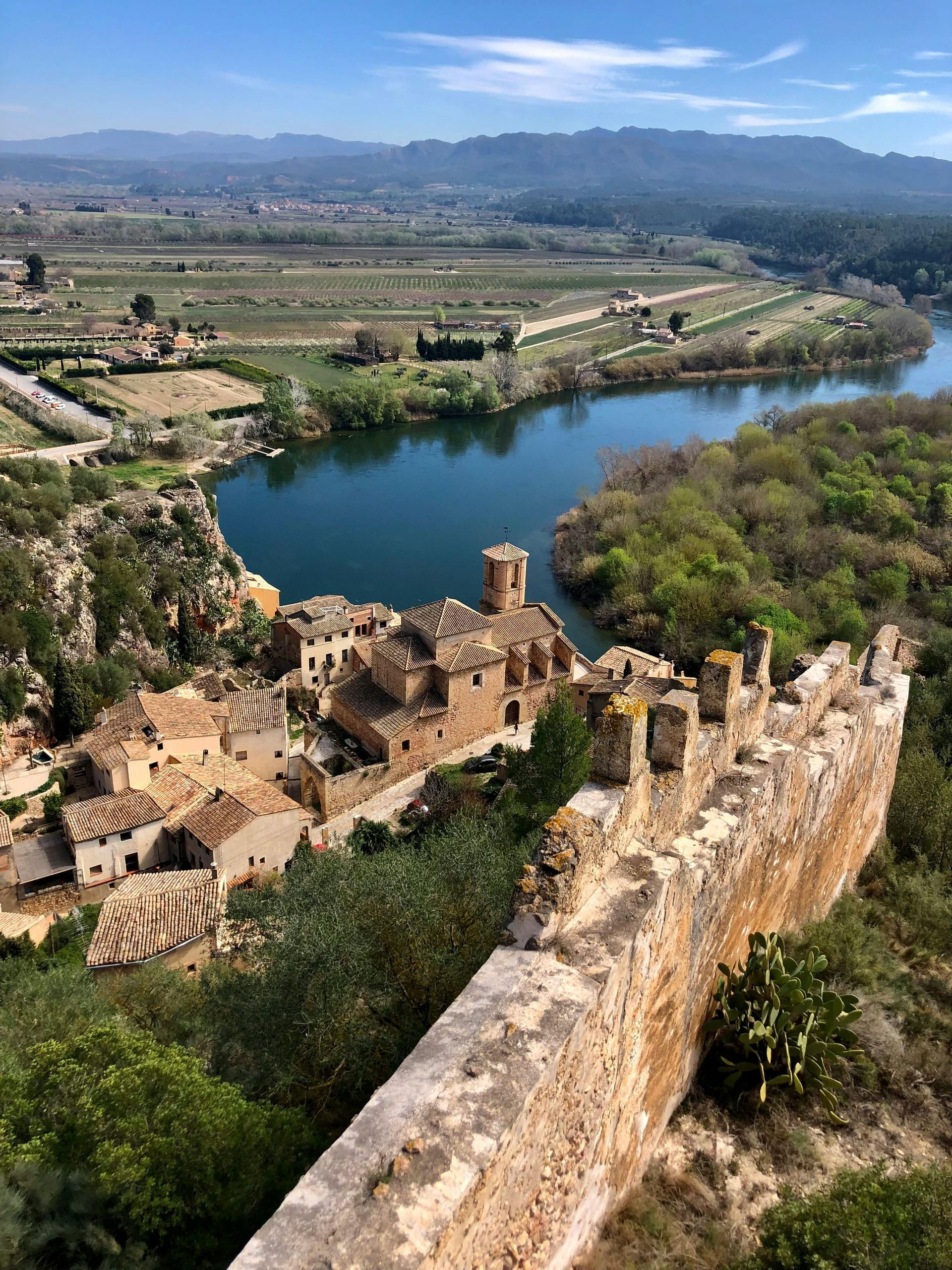 Ruinous stone buildings and a river winding through a valley, seen from a weathered stone wall. Camino de Santiago