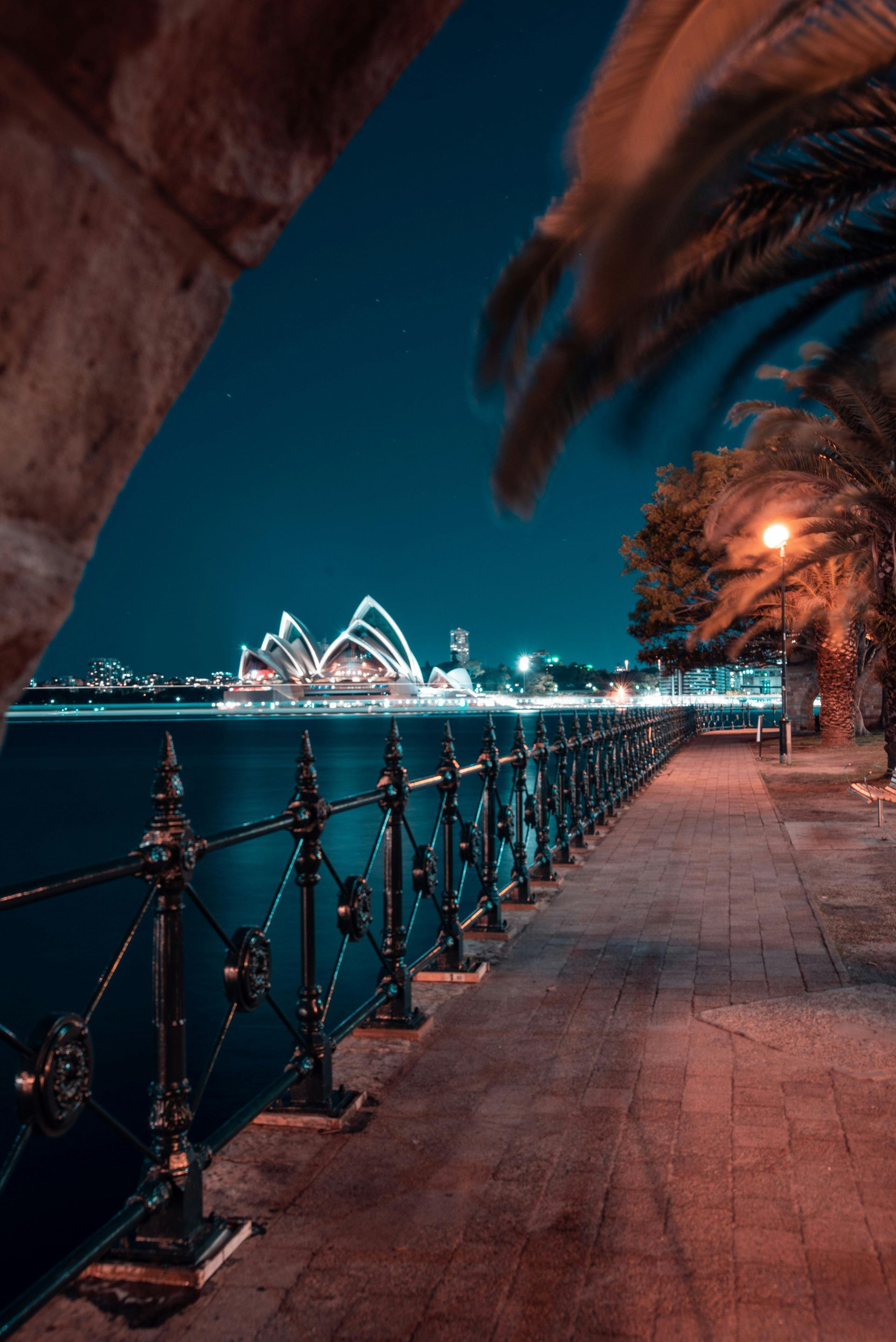 Sydney Opera House at night, viewed through an archway, along a waterfront promenade with decorative railing and palm trees. Gay City Breaks in Sydney