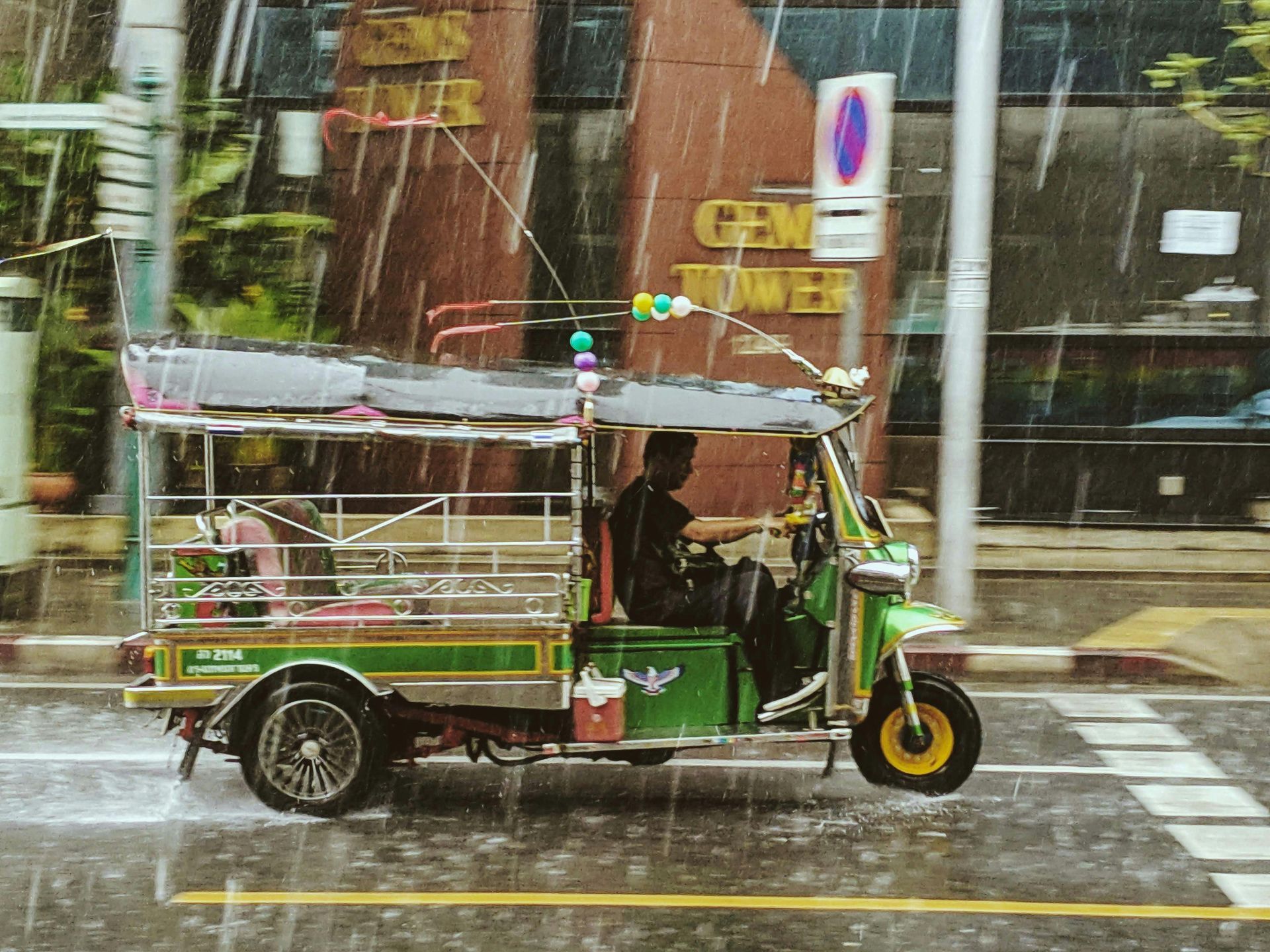 A green tuk-tuk driving in the rain on a city street, with a driver visible. Thailand, Bangkok