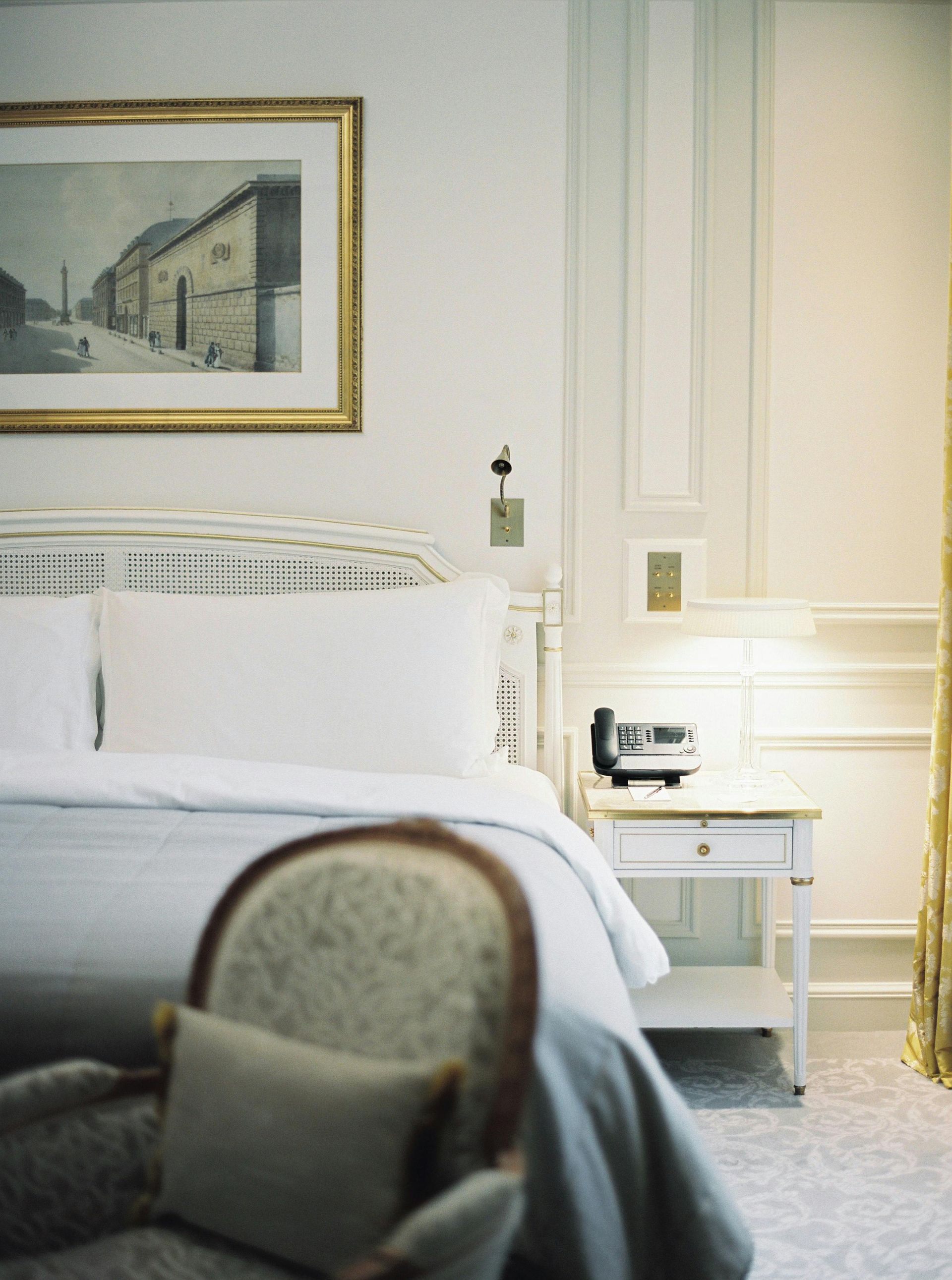 A hotel bedroom features a bed with white linens, a framed print, and an ornate armchair in the foreground.