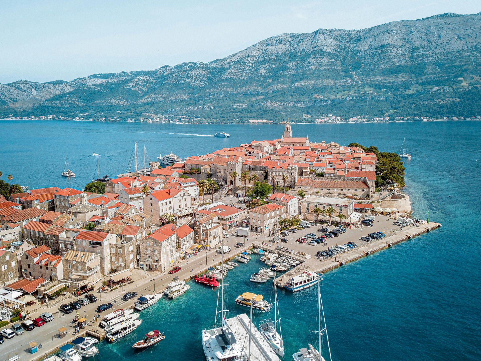 Aerial view of Korčula, Croatia, with its stone buildings, red-tiled roofs, harbor, and boats against a mountain backdrop.