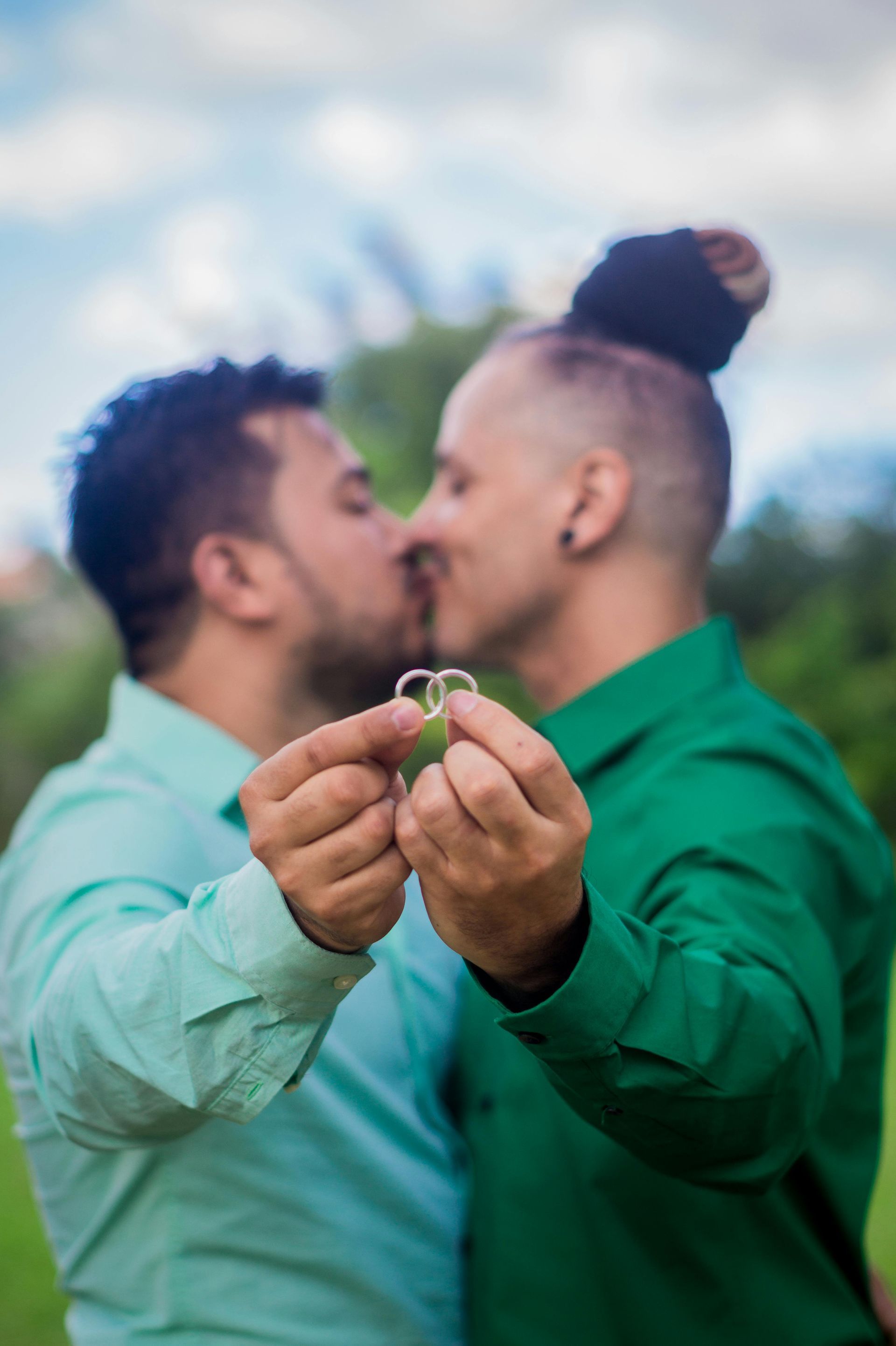 Two men kissing, holding rings. One in green, the other in turquoise, outdoors.