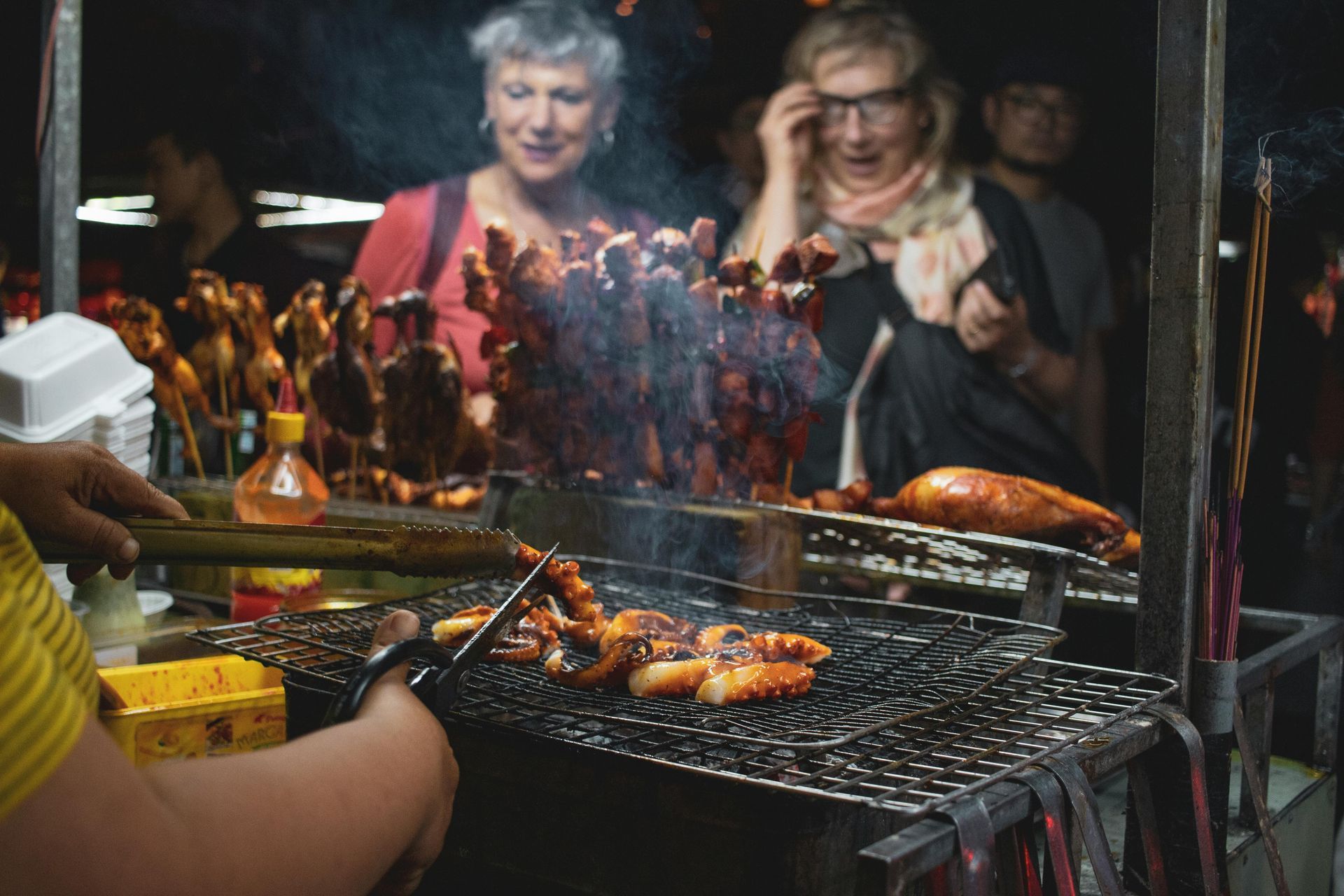 Street food vendor grilling skewers of squid, with two women looking on, lit by smoke and dim lights.