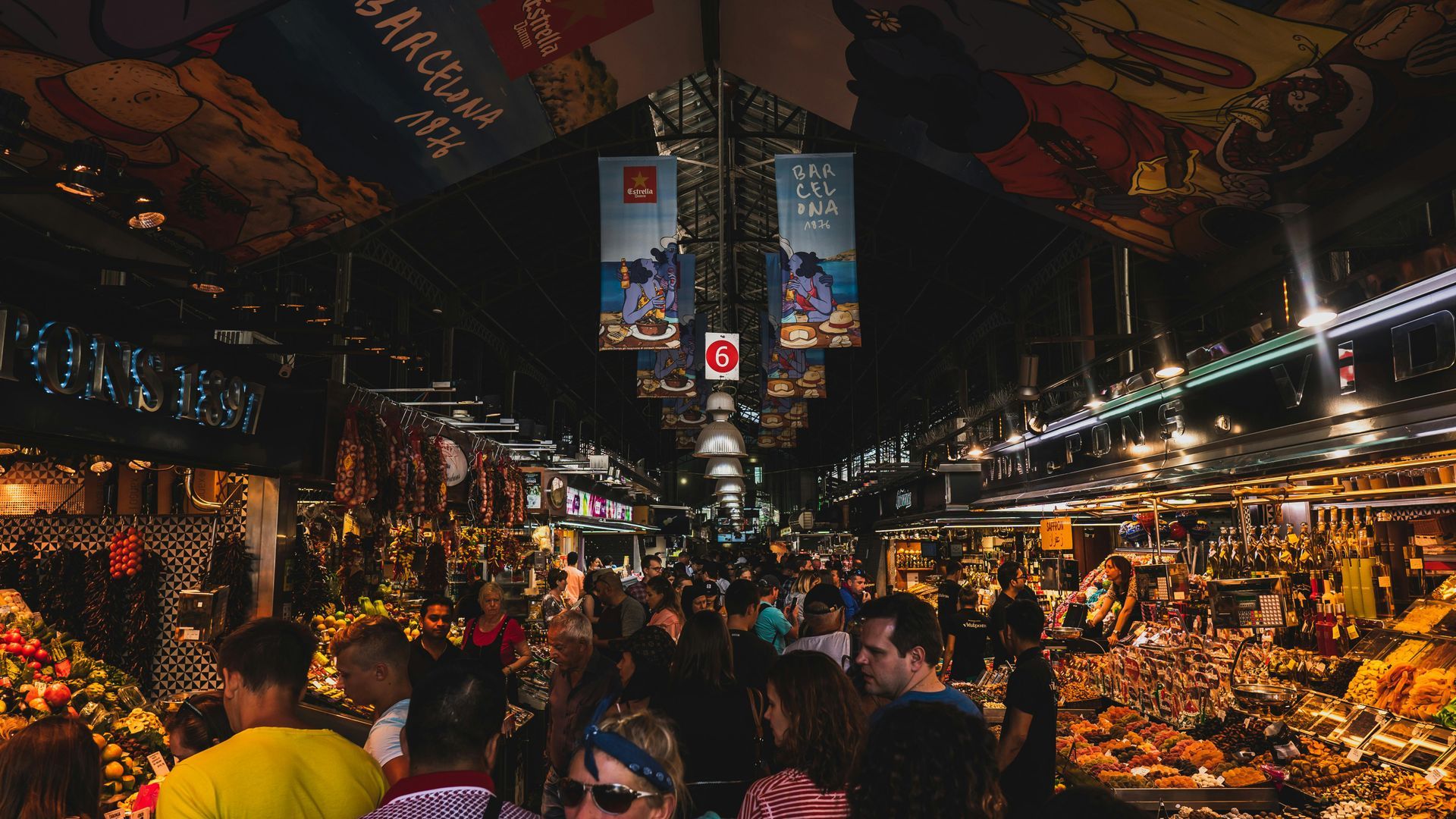 Crowded marketplace at night. People browse stalls under colorful banners and lights.
