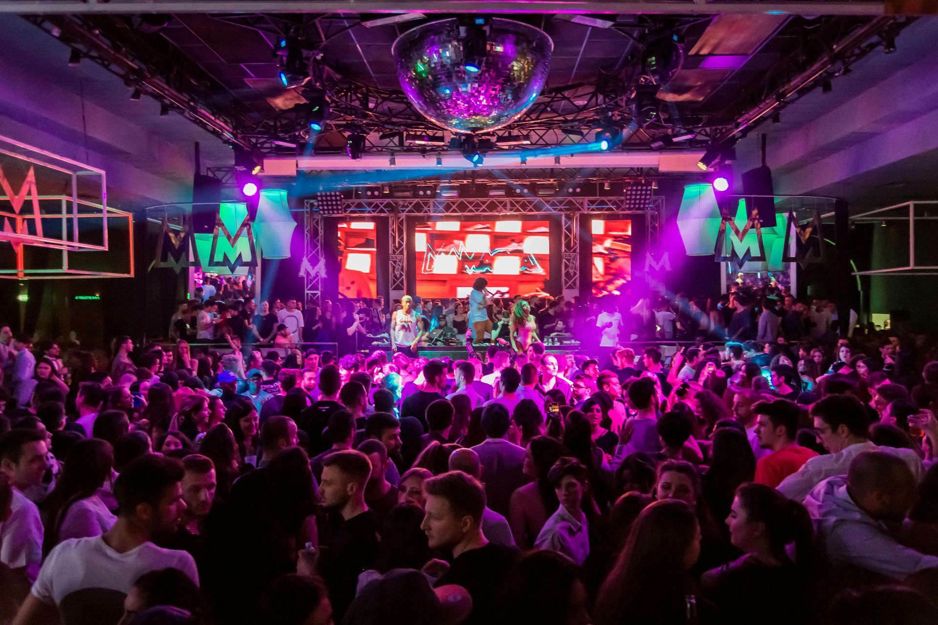 A crowded nightclub with purple lighting, a disco ball, and a DJ booth illuminated by large video screens.