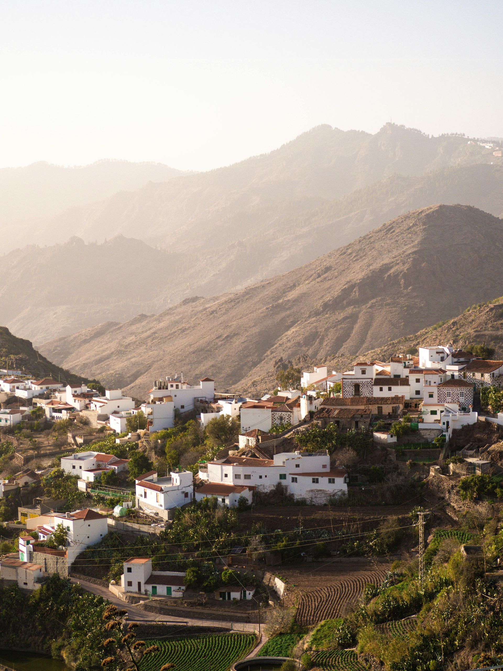 White buildings nestled in a hillside, with brown mountains in the background. A warm, sunny haze filters the landscape.Gran Canaria,