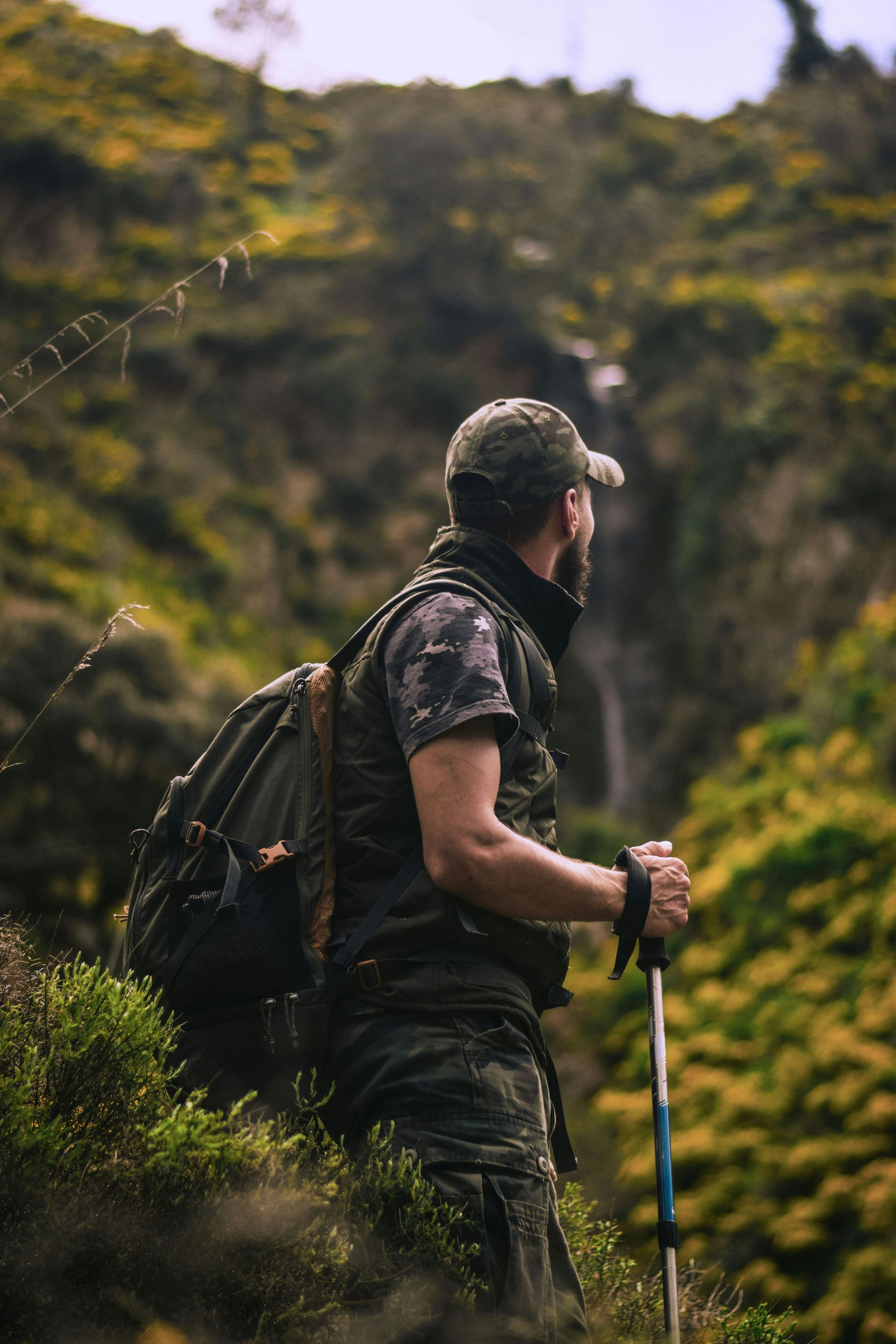 Man in camouflage hiking, backpack and pole, looking up at a waterfall in a green, mountainous area.