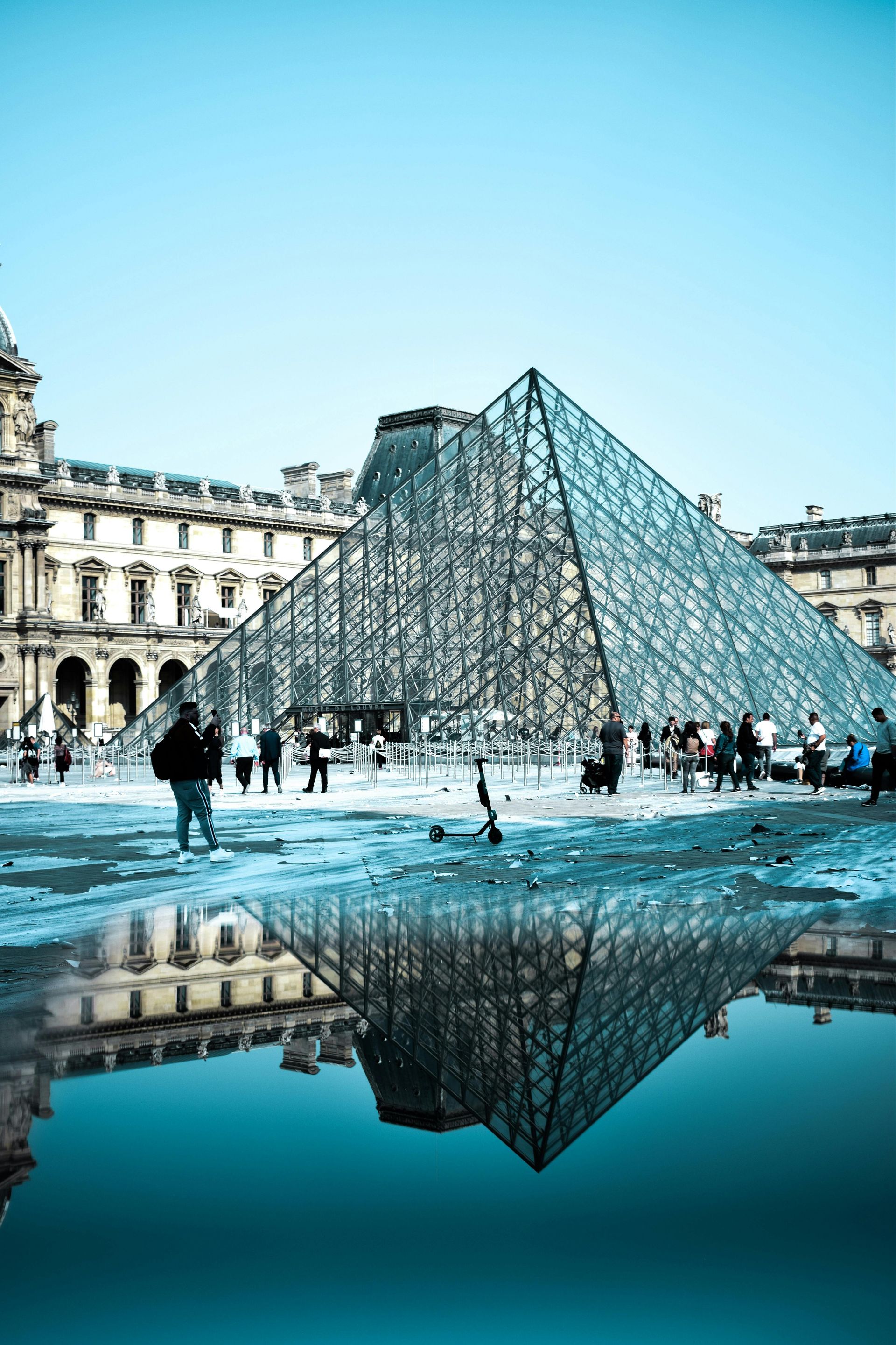 Louvre Pyramid reflected in a puddle; Paris, France. Blue sky, glass structure, people nearby.