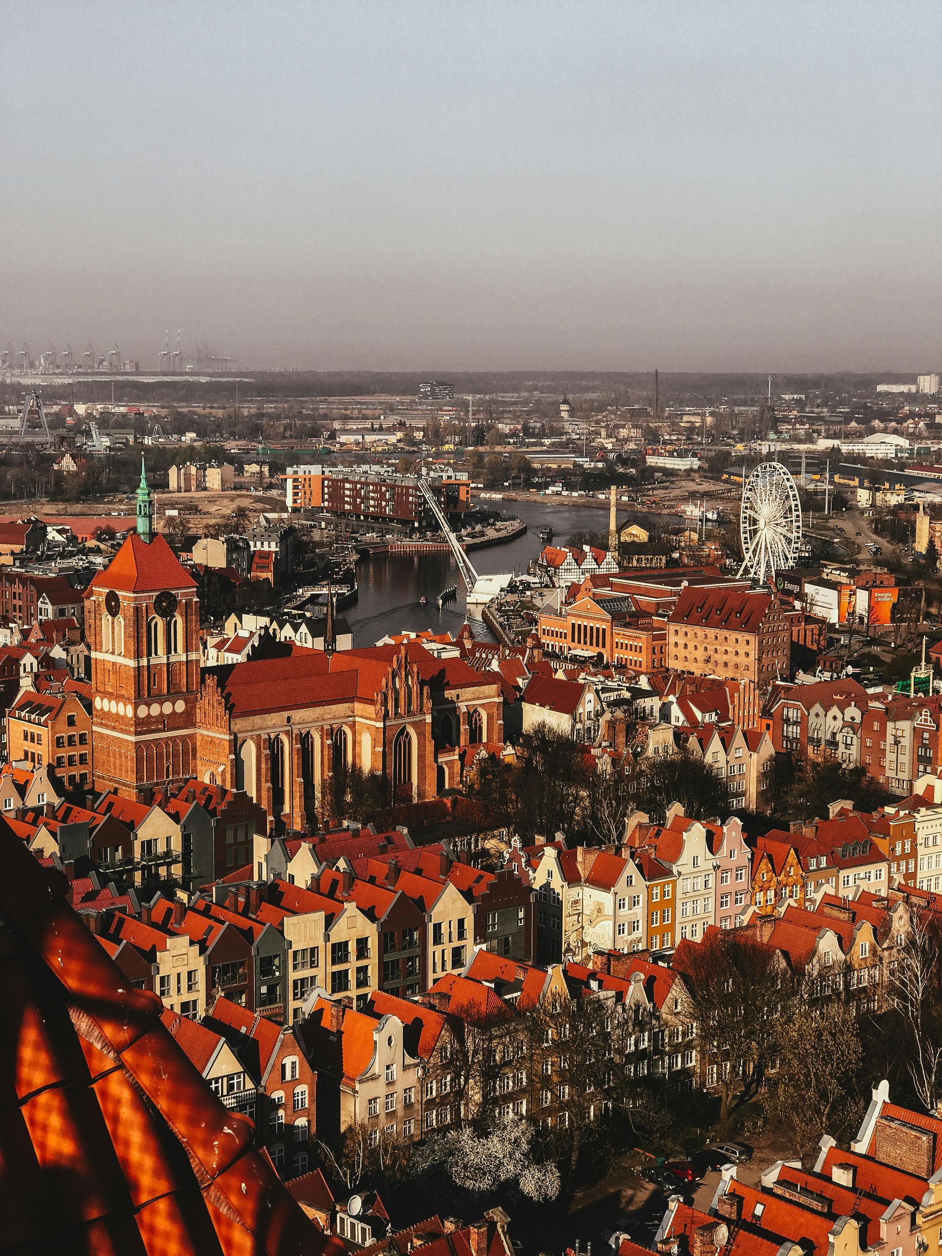 Aerial view of Gdansk, Poland, showcasing red-roofed buildings and a river winding through the city under a hazy sky.
