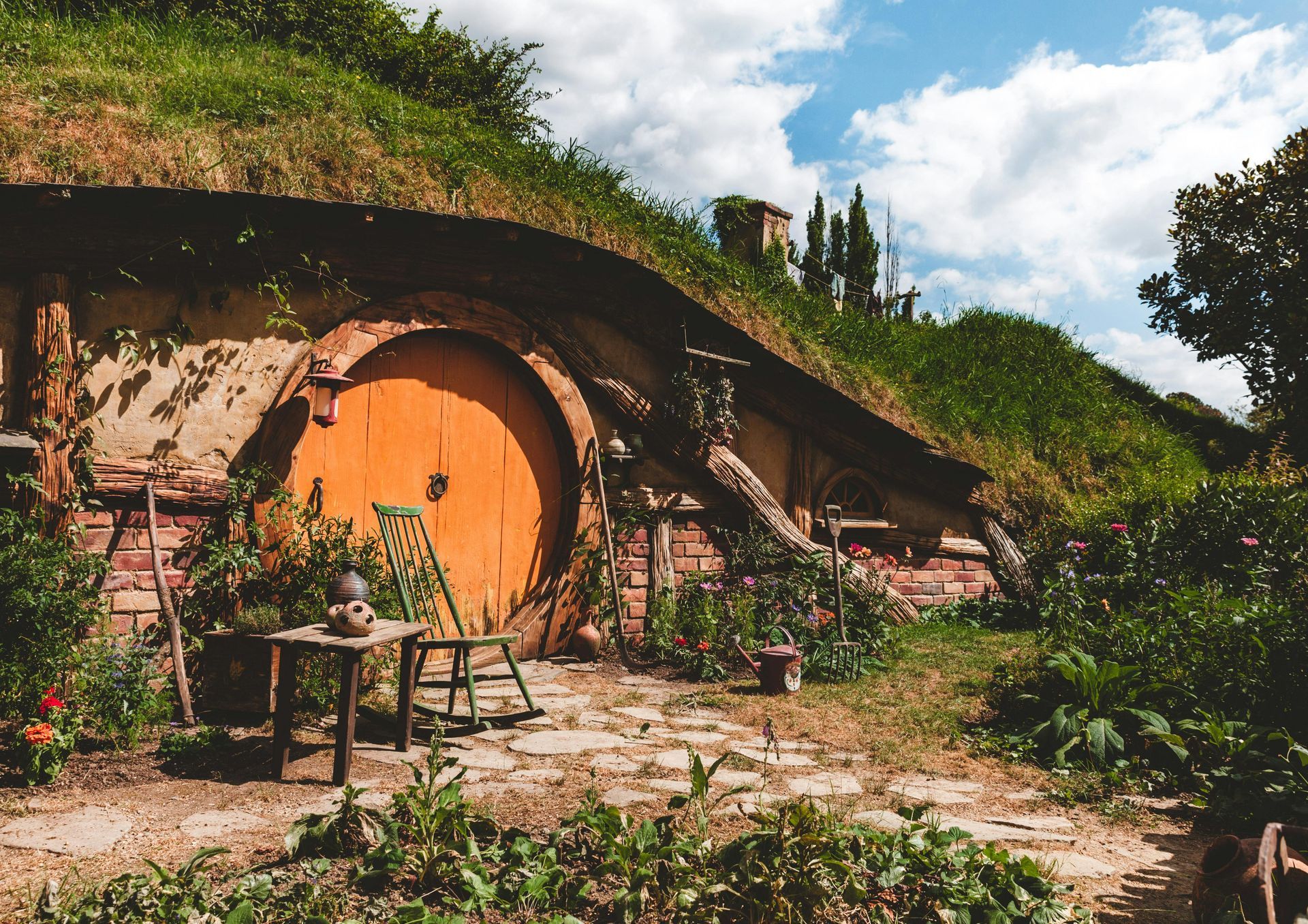 Hobbit hole with round orange door, green grassy roof, small chair, and garden. New Zealand