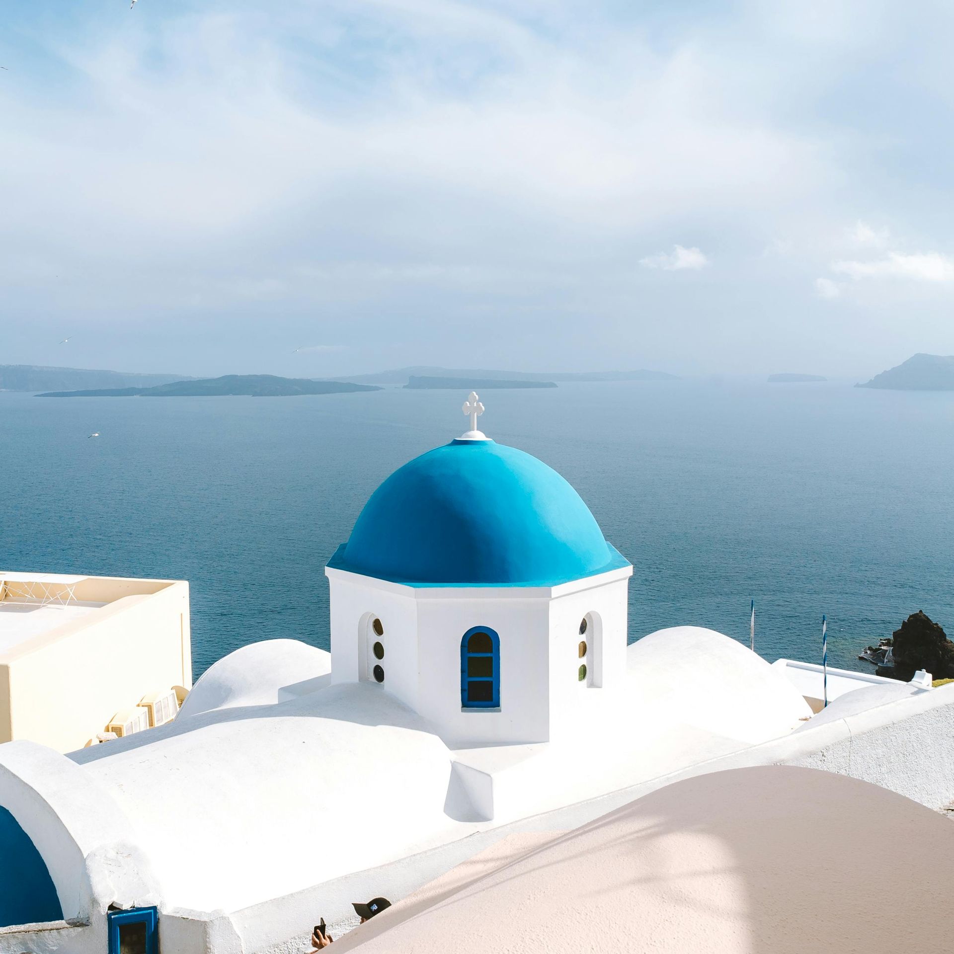White buildings with blue-domed church against a backdrop of the Aegean Sea under a cloudy sky in Santorini, Greece.