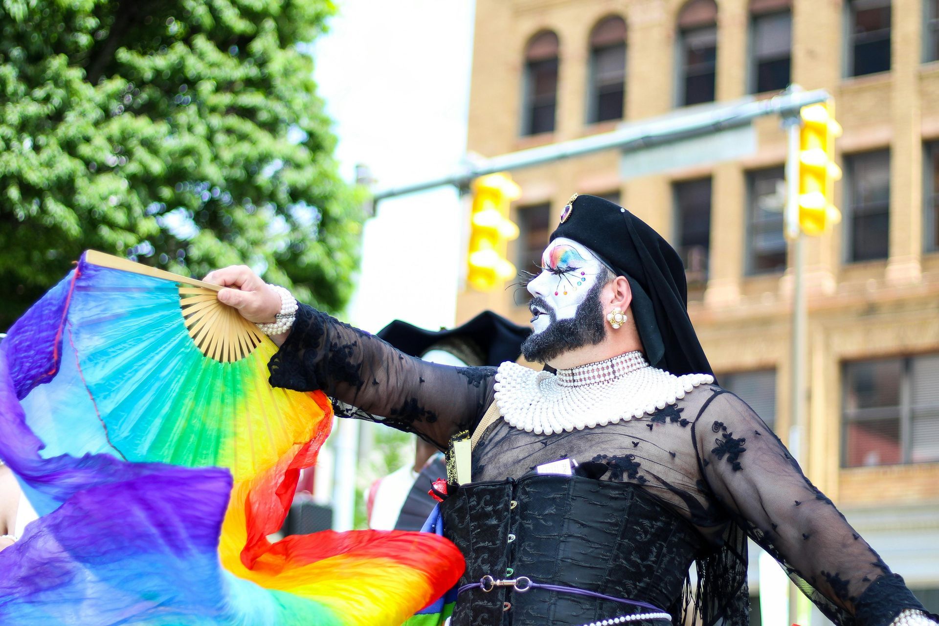 Person in drag with rainbow fan at Pride parade, wearing black lace and makeup, street setting.