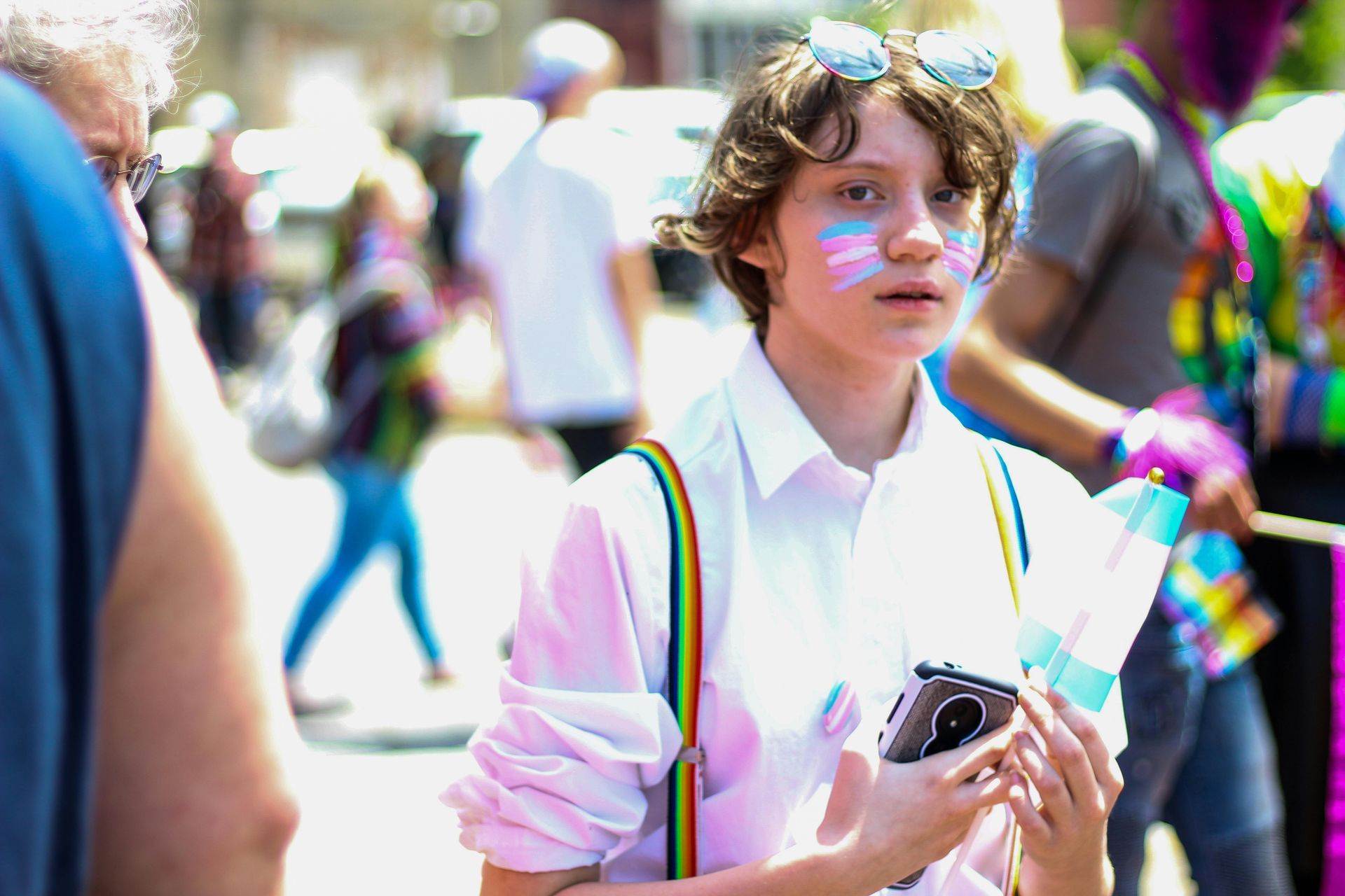 Person at a pride event with face paint and trans flag. Wearing white shirt and rainbow suspenders.