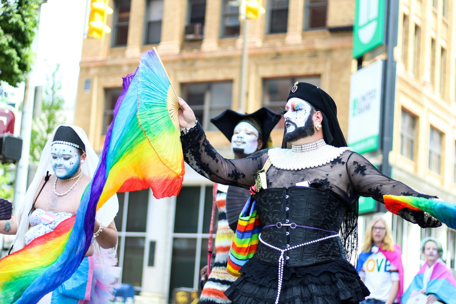 People in drag at a Pride parade, waving rainbow flags. Downtown street setting.