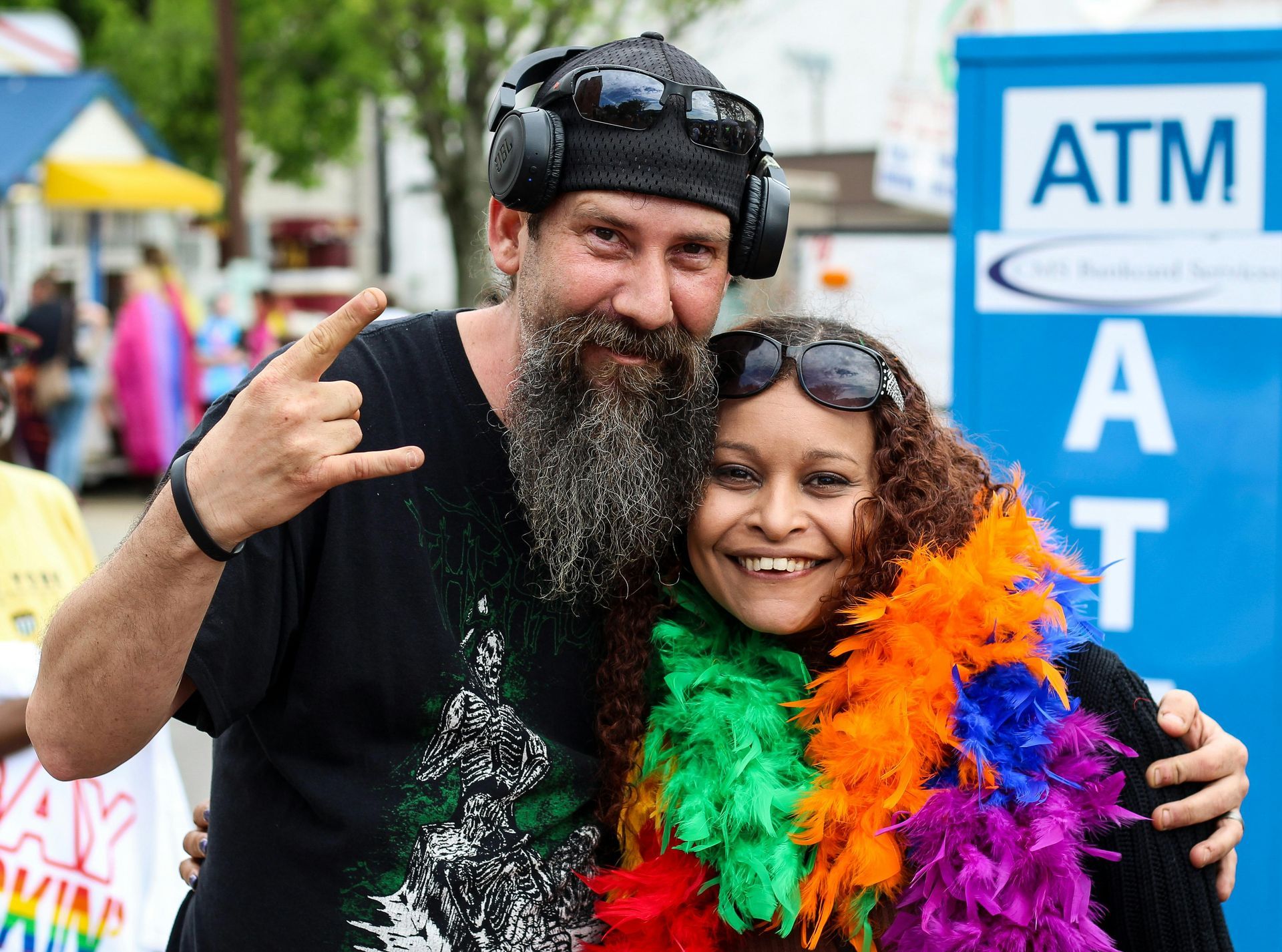 Man with beard and woman with rainbow boa pose by an ATM.