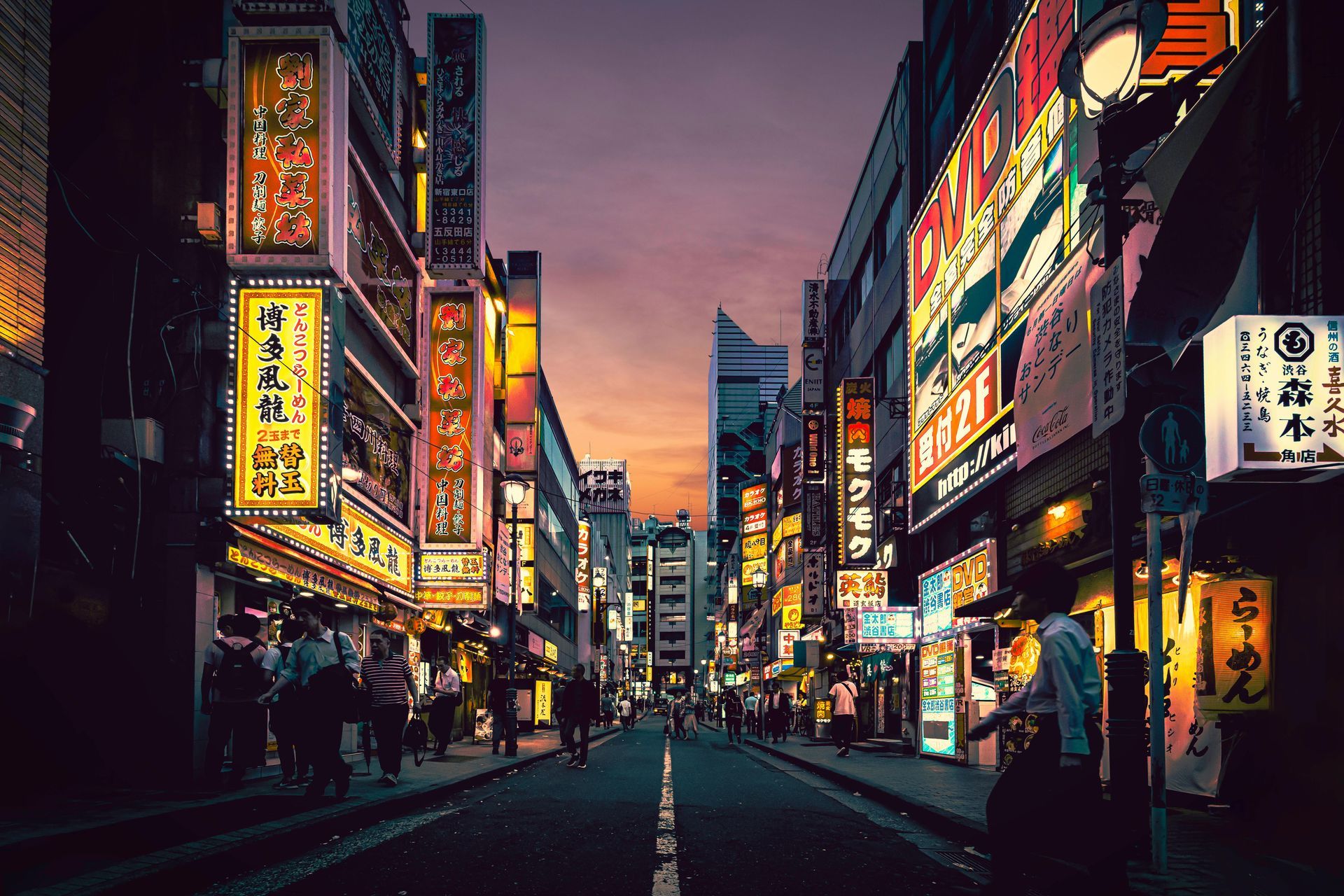 A vibrant street in a city at dusk, lined with glowing signs and shops. People stroll along the road as the sky fades to purple.