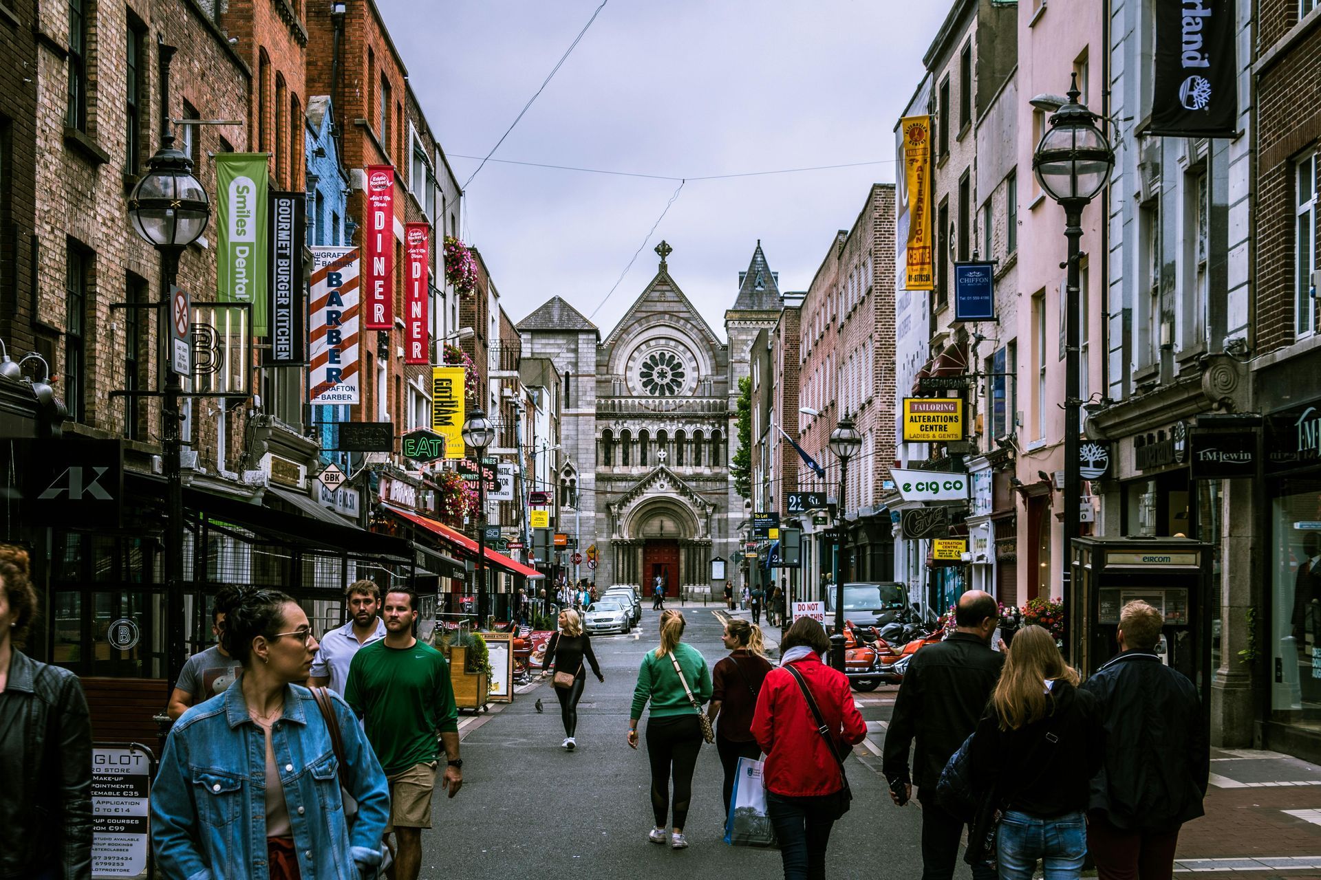 Pedestrians walk down a crowded city street toward an ornate stone church at the end of a corridor of buildings.