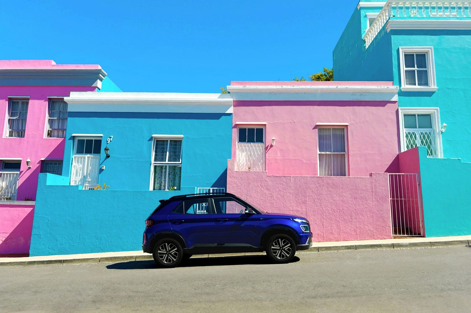 Blue SUV parked in front of colorful pink and turquoise buildings on a sunny day.