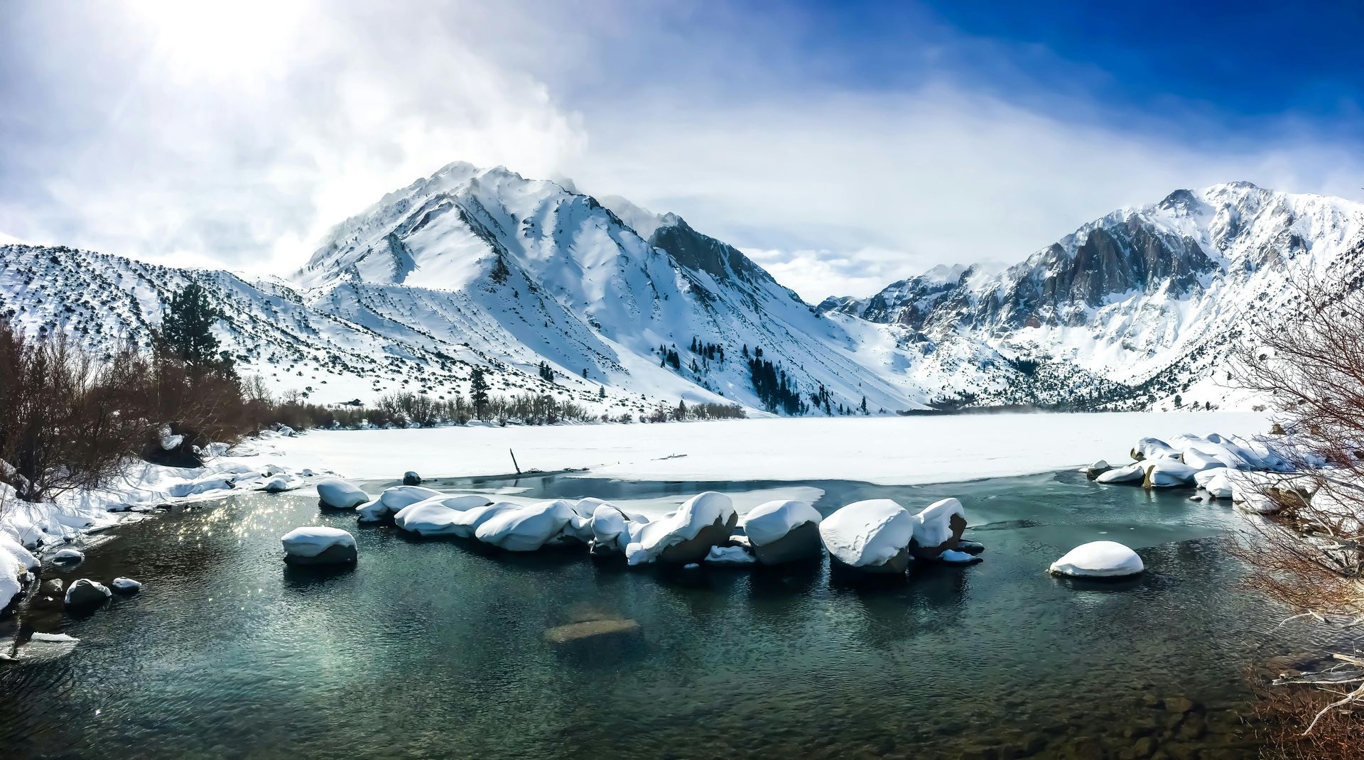 Snow-covered mountains frame a partially frozen lake with snow-dusted rocks. Blue sky, winter landscape.