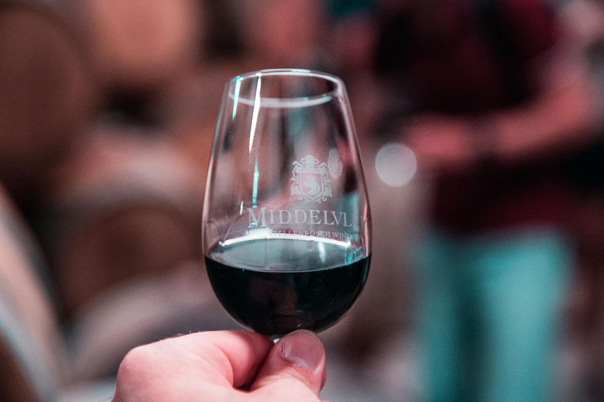 Hand holding a wine glass with dark red liquid; blurred background of barrels. Cape Winelands,