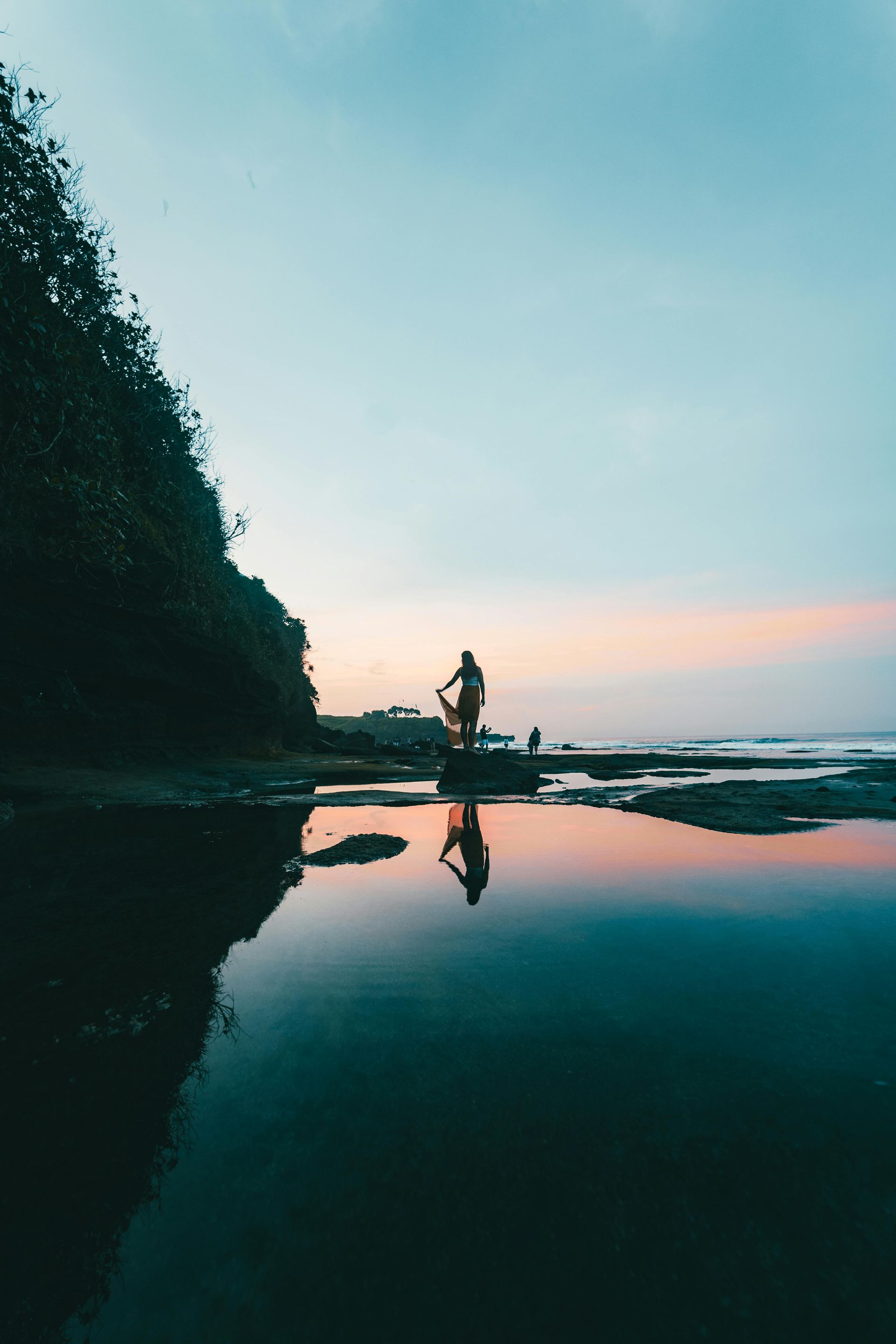 Person on a rock at the beach, reflected in still water. Twilight sky, dark foliage. Bali