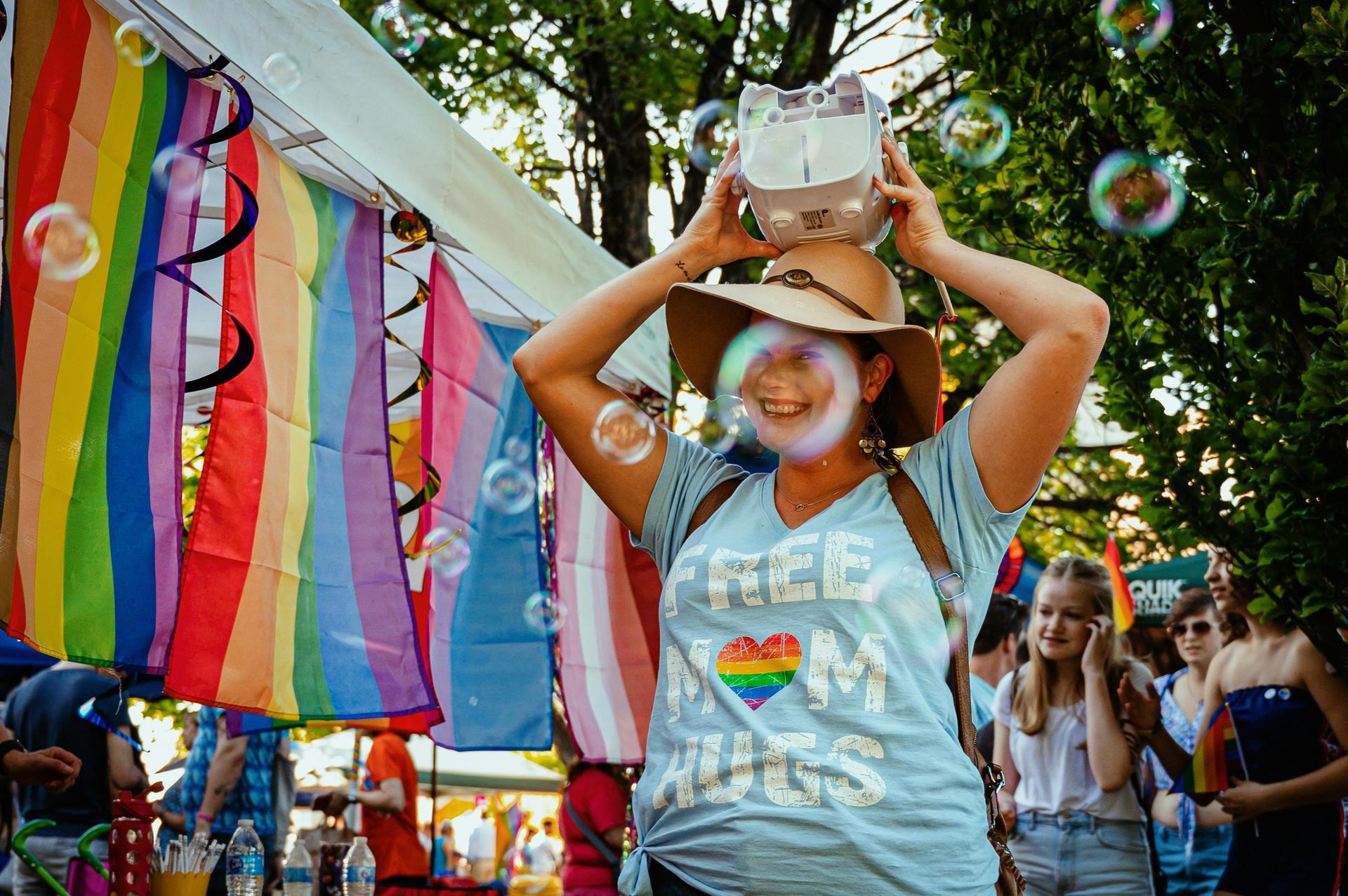 A person in a sun hat holding a VR headset above their head, surrounded by rainbow flags and soap bubbles at an event.
