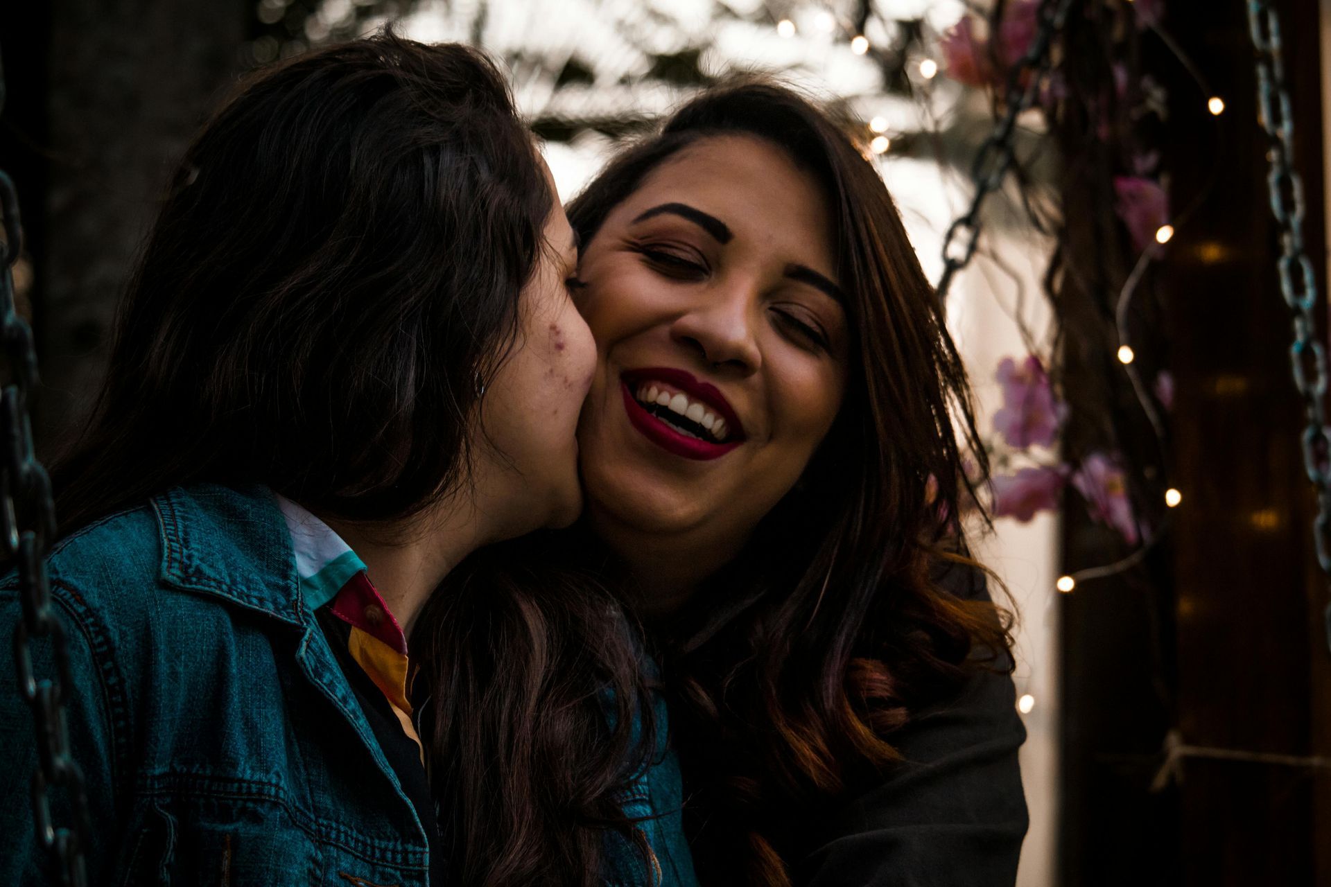 Woman kissing another woman's cheek, both smiling. Fairy lights and floral accents in the background.