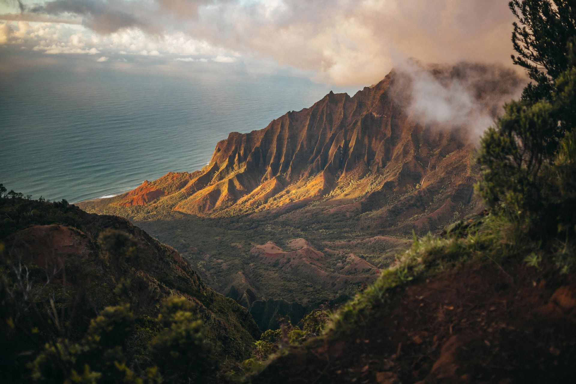 Mountains rising from the ocean, bathed in sunlight. Lush green valley with cloudy sky. Kalalau Trail,