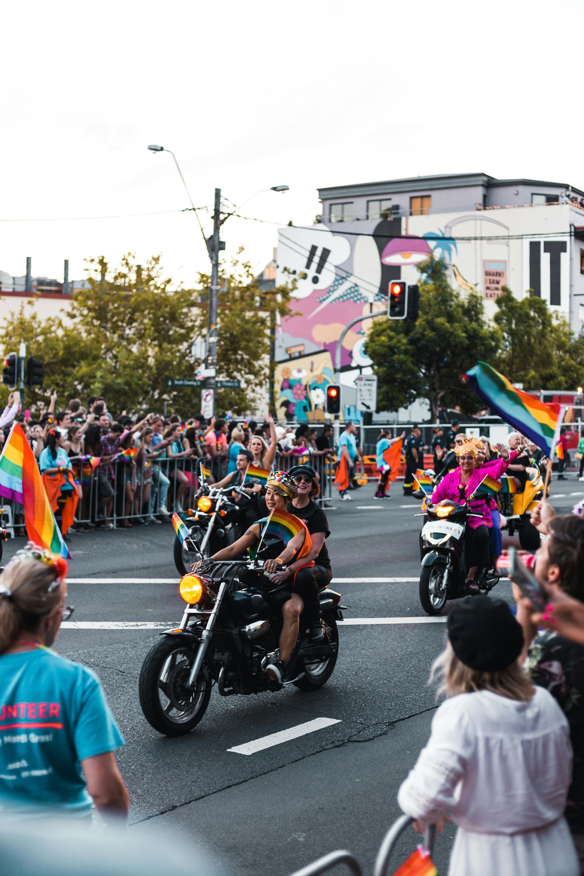 A parade with people on motorcycles, rainbow flags, and spectators. A colorful mural is visible in the background. Sydney Mardi Gras
