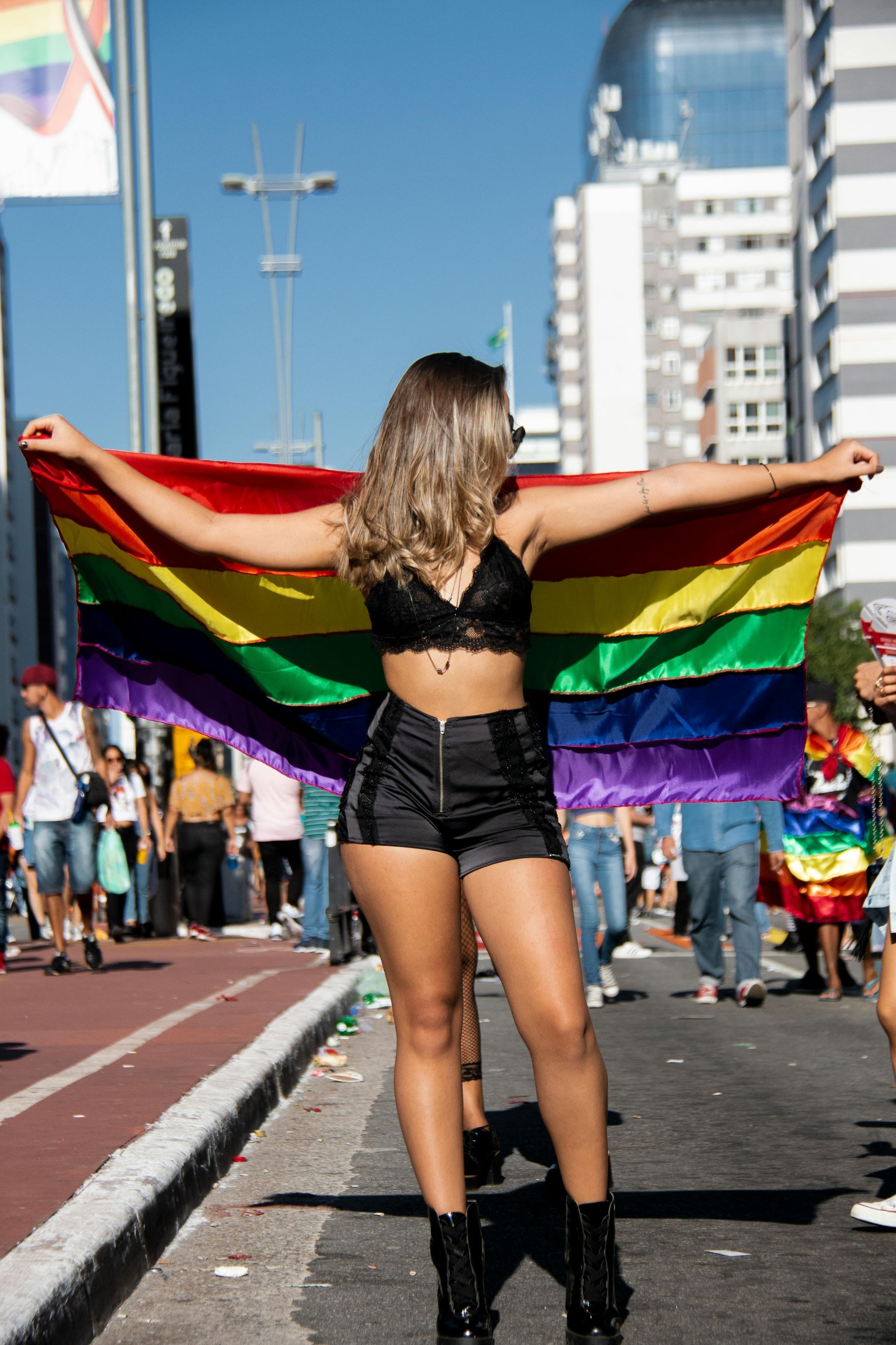 A person stands in a city street holding a large rainbow flag outstretched behind their back at a pride parade.
