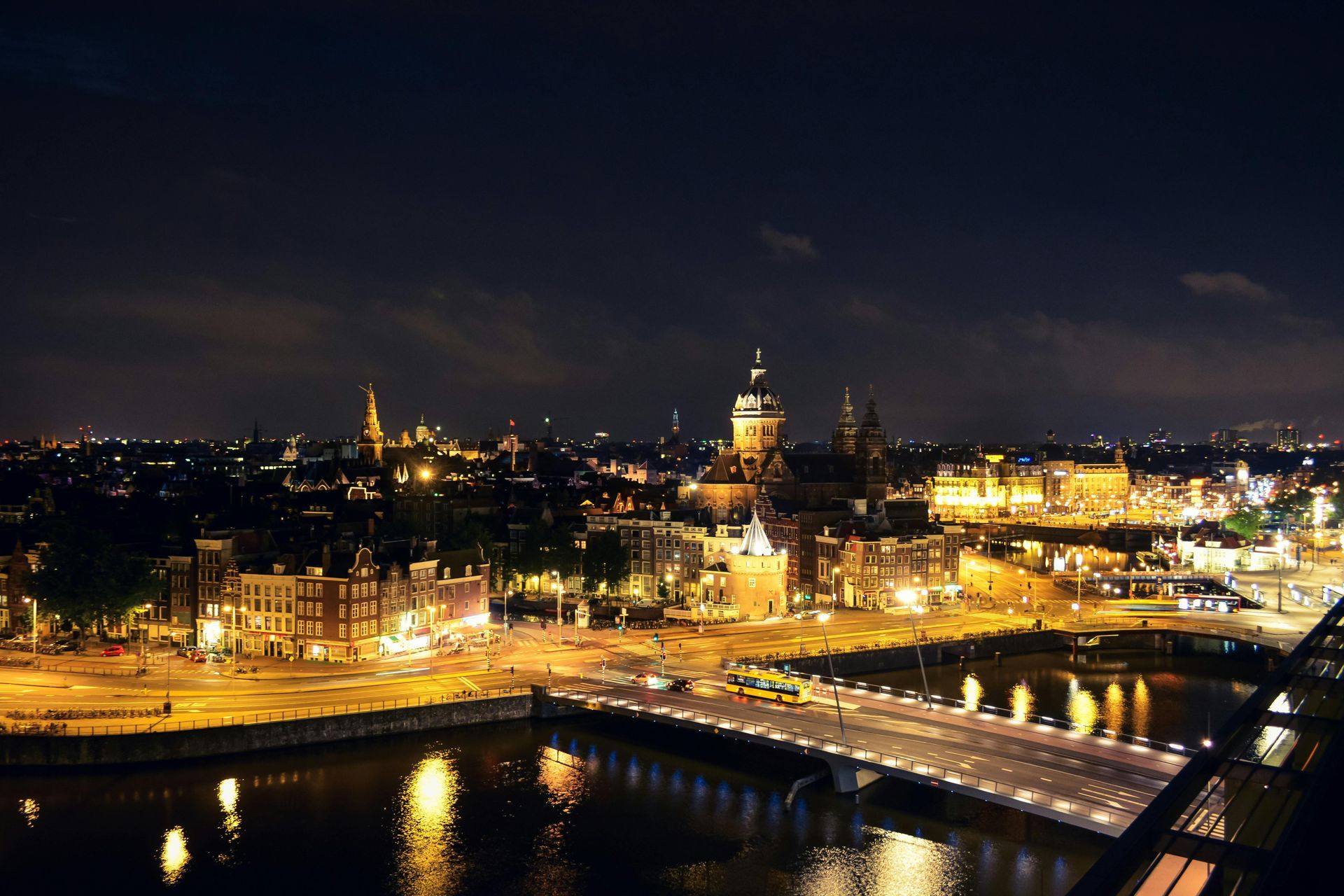 A night view of Amsterdam, featuring illuminated buildings, the Saint Nicholas Basilica, and a bridge crossing the water.