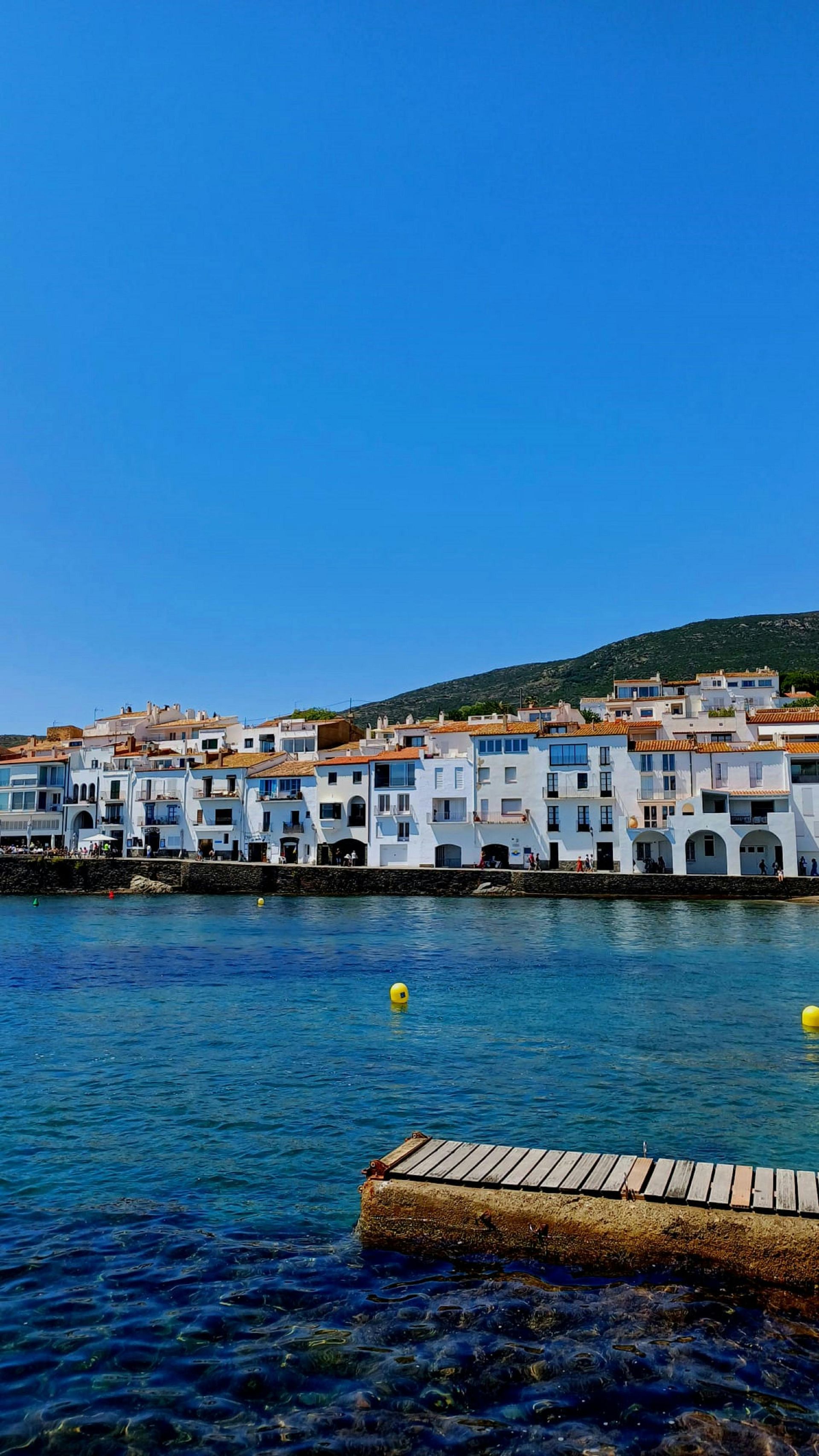 White buildings line a shore, with blue water and sky. Cadaqués, Spain