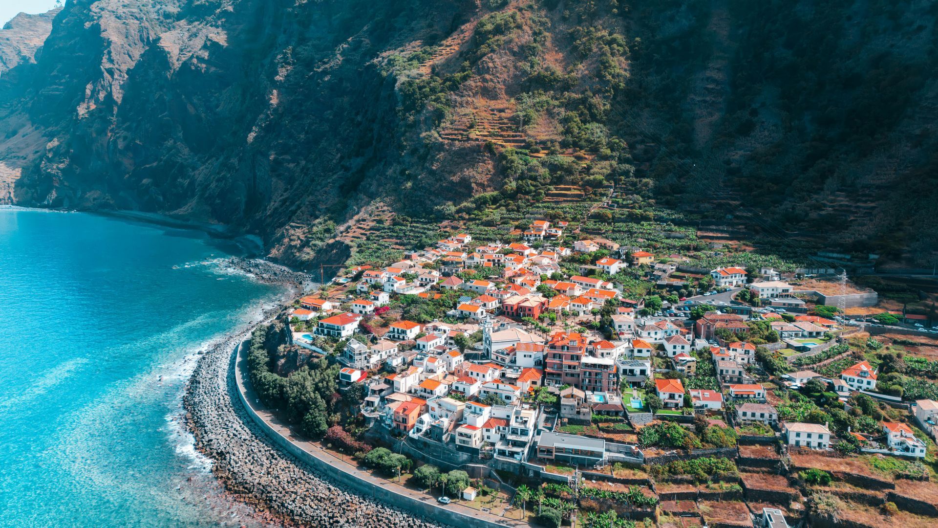 Coastal village nestled at the base of a mountain, with blue ocean and orange rooftops. Madiera