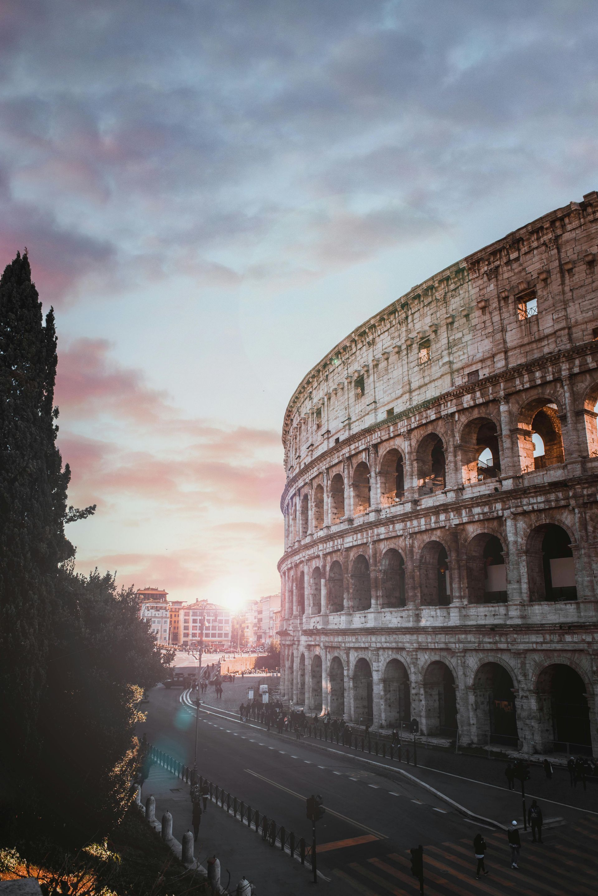 The Colosseum in Rome, partially lit by a sunset with pink and orange hues. A road and trees are in view.