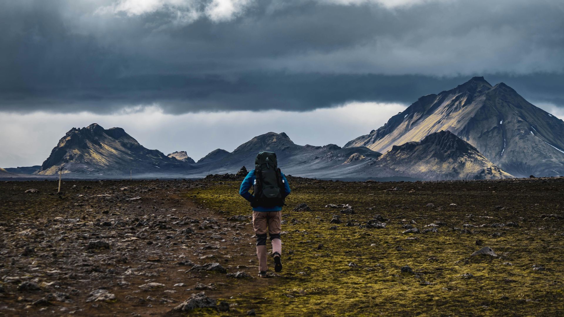 Person backpacking across a barren landscape towards mountains under a stormy sky.
