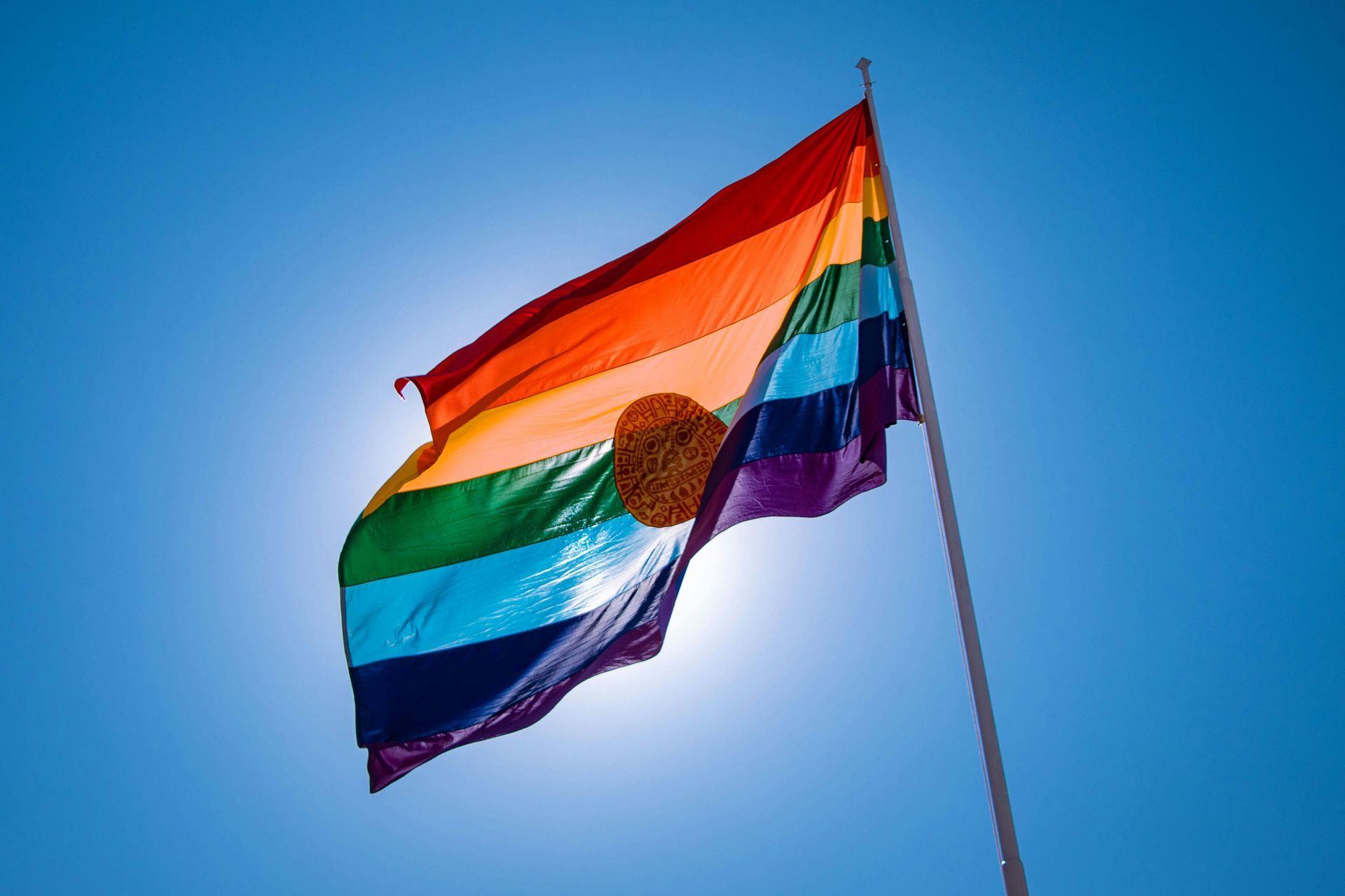 Rainbow flag waving against a clear blue sky, sun shining through.