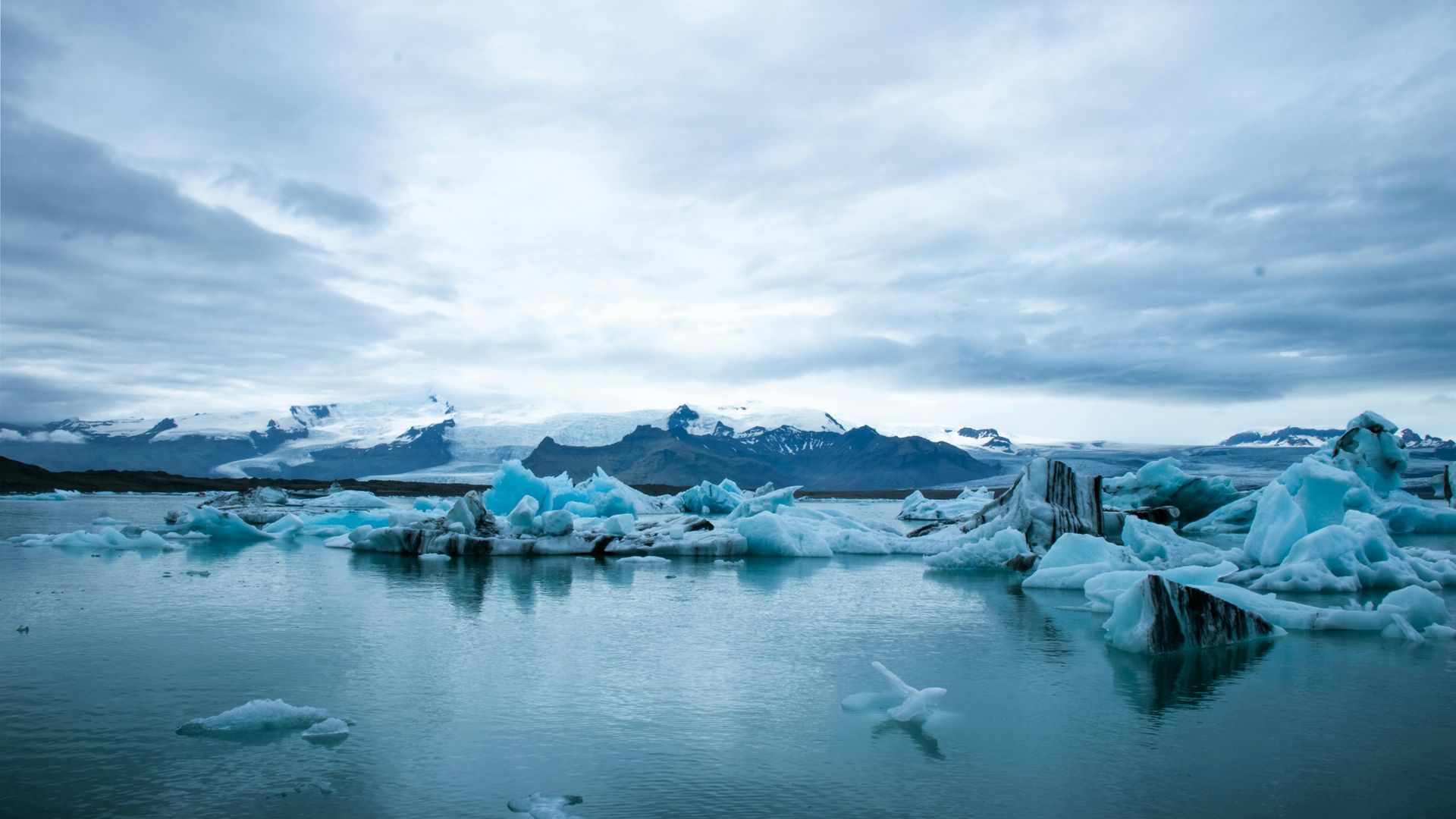 Glacial lagoon with blue icebergs, mountains, and overcast sky.