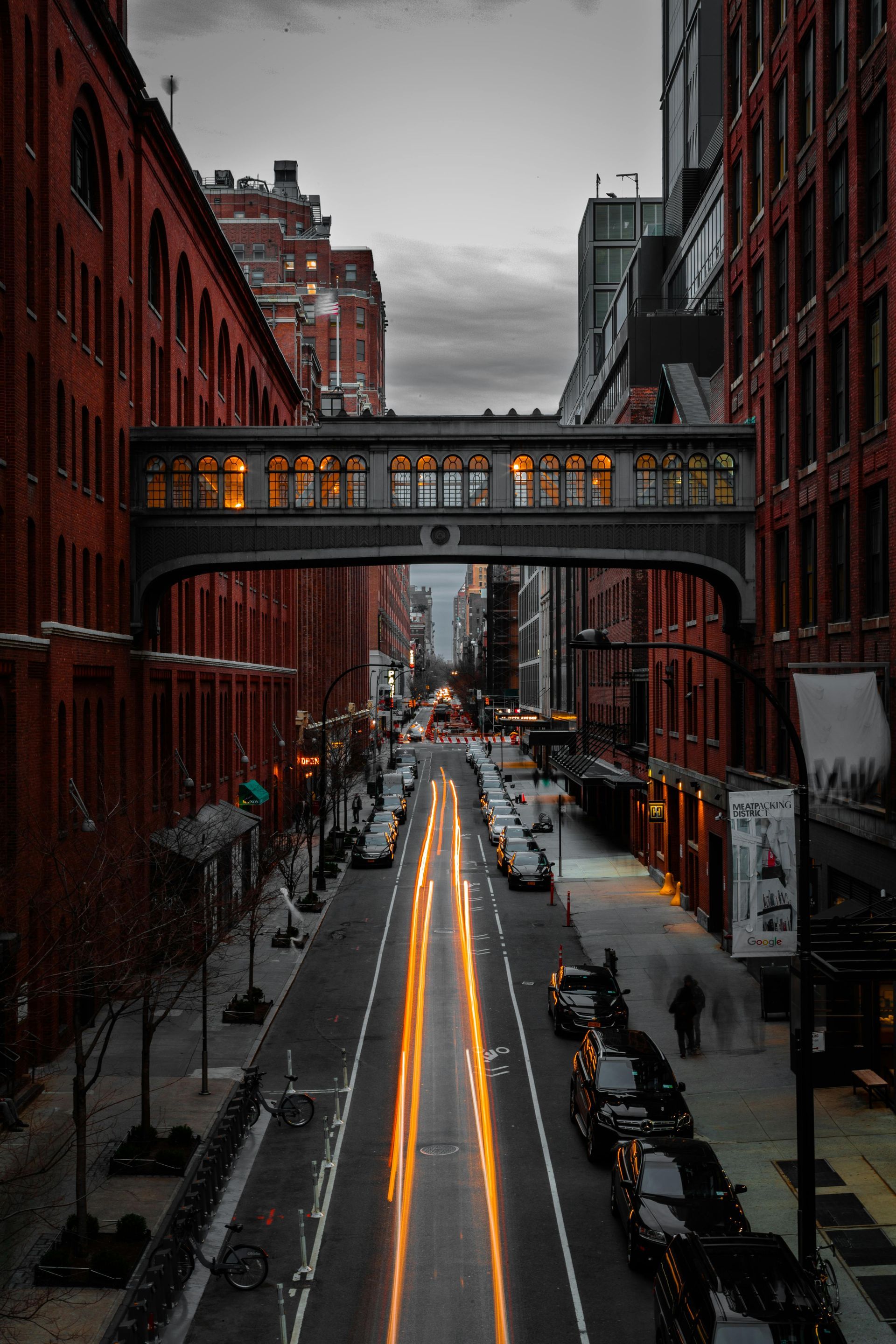 City street at dusk, lined with red brick buildings and a skybridge, streaked with car light trails. Chelsea – New York City