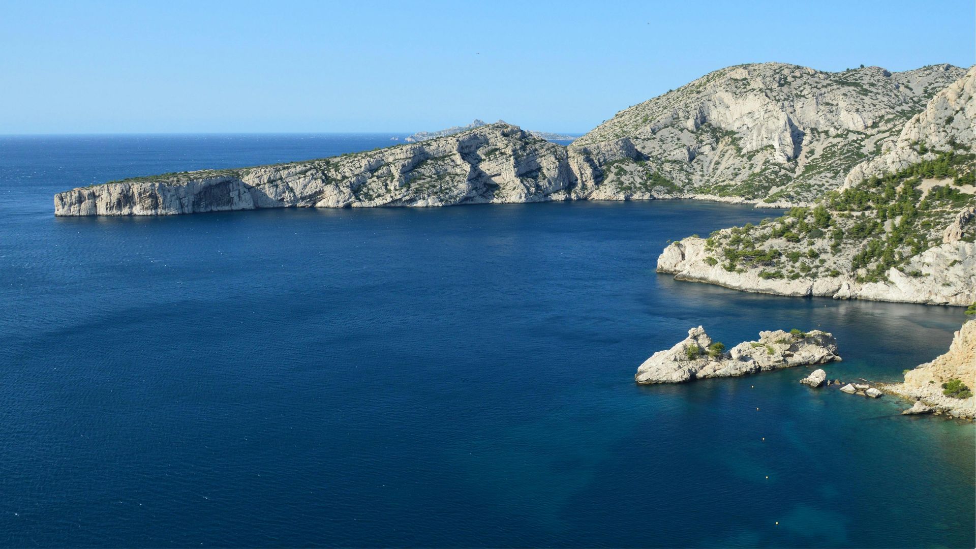 Rocky cliffs overlooking a deep blue Mediterranean cove under a clear blue sky.