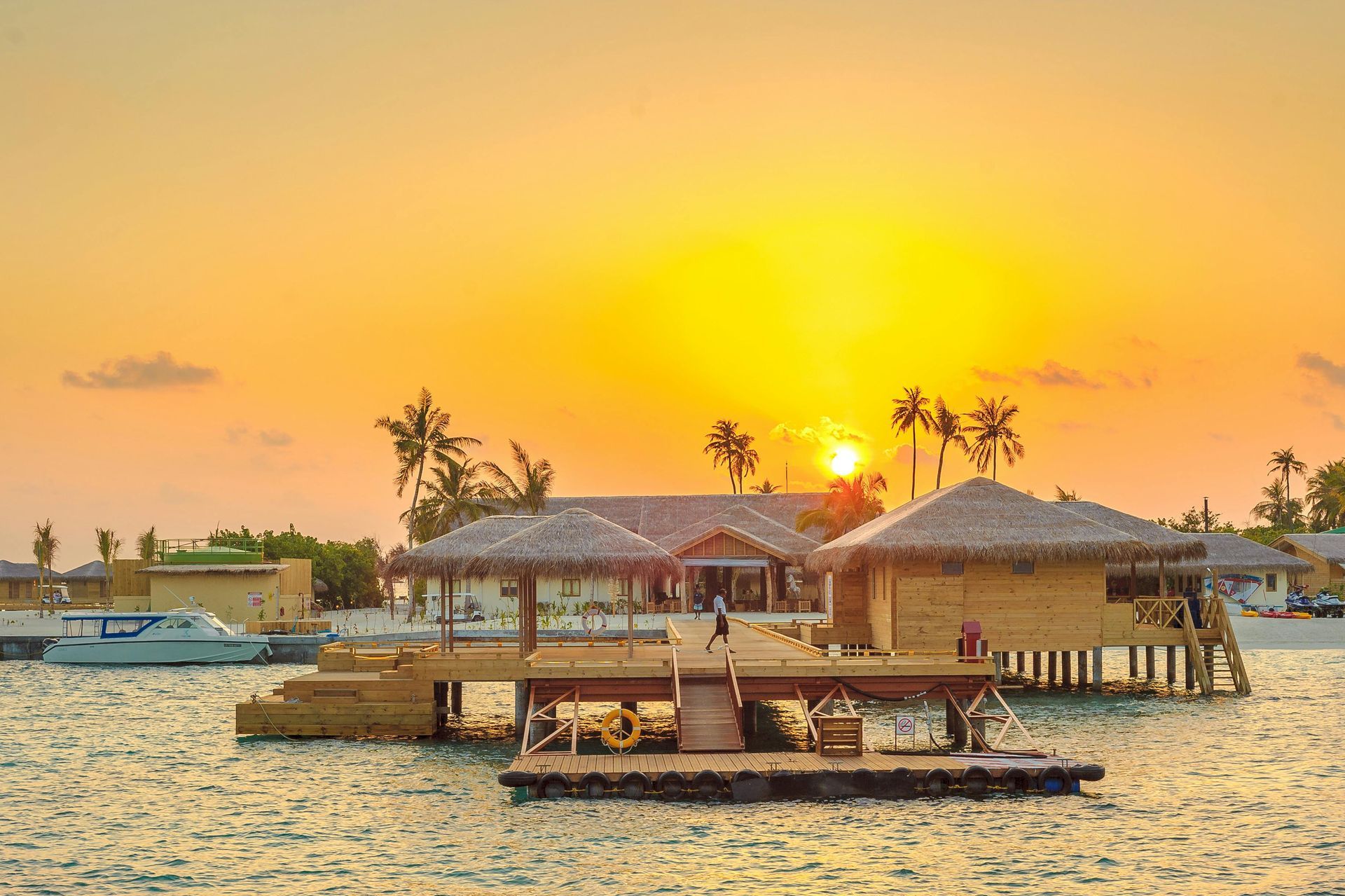 Sunset over a pier with thatched-roof structures on water; a boat is docked, and palm trees are in the background.