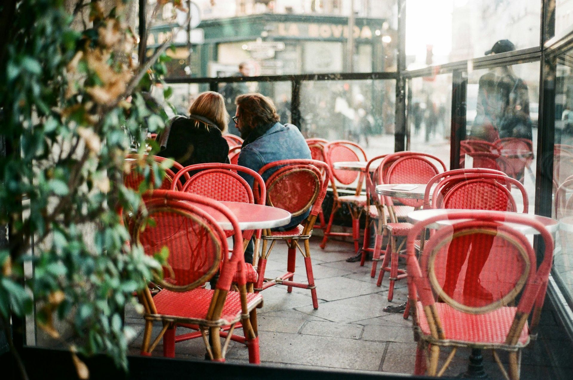 Red chairs at an outdoor cafe in Paris, two people seated and talking; green plants in foreground.