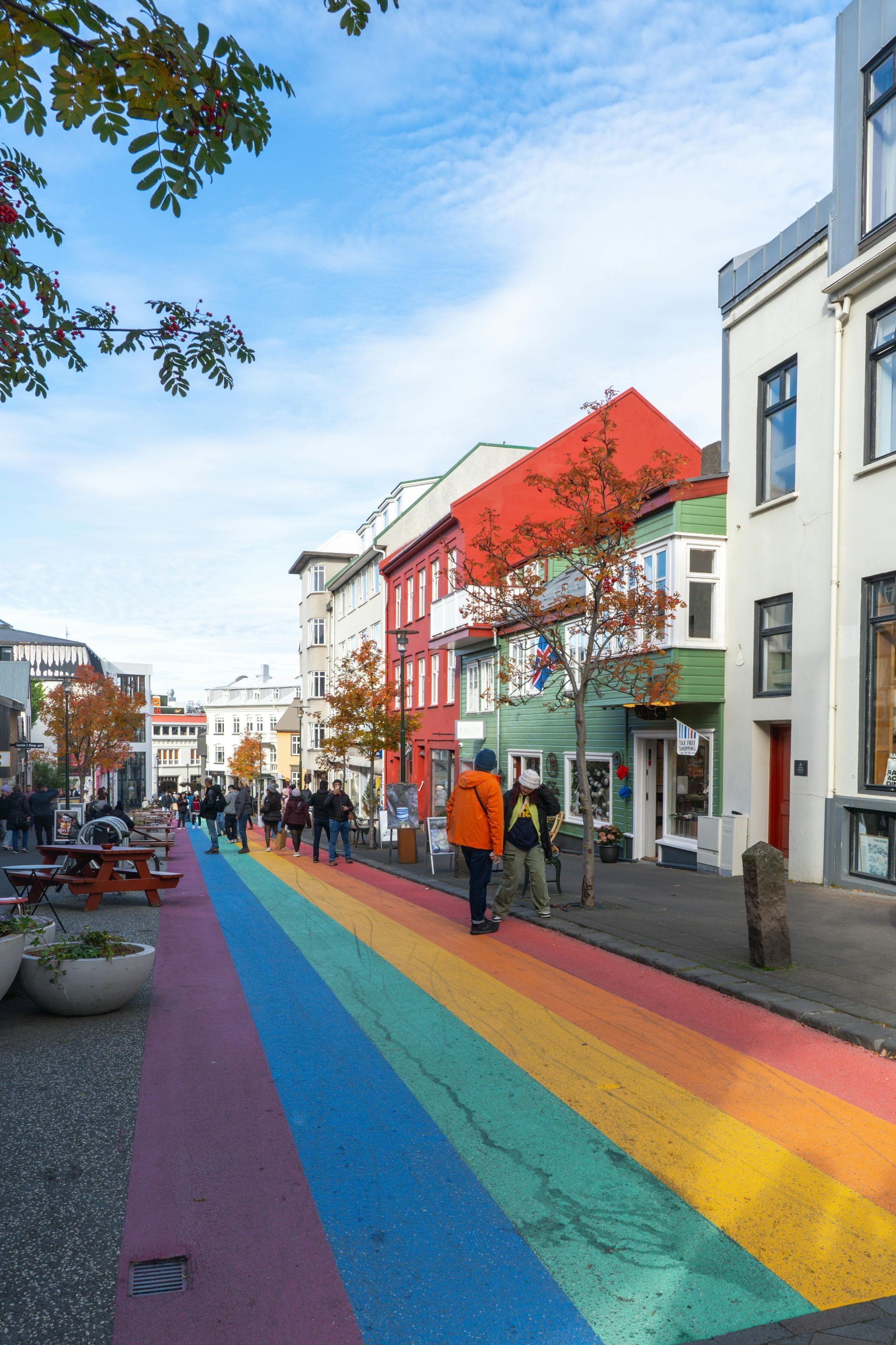 Rainbow-painted street in Reykjavik, Iceland, with pedestrians walking past colorful buildings on a sunny day.