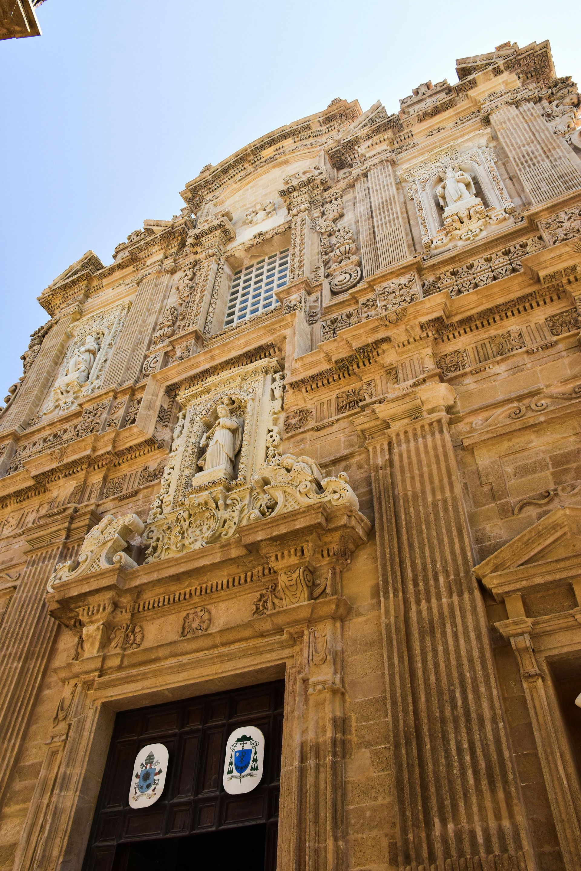 Ornate stone church facade with sculptures and dark wooden doors against a blue sky. Lecce
