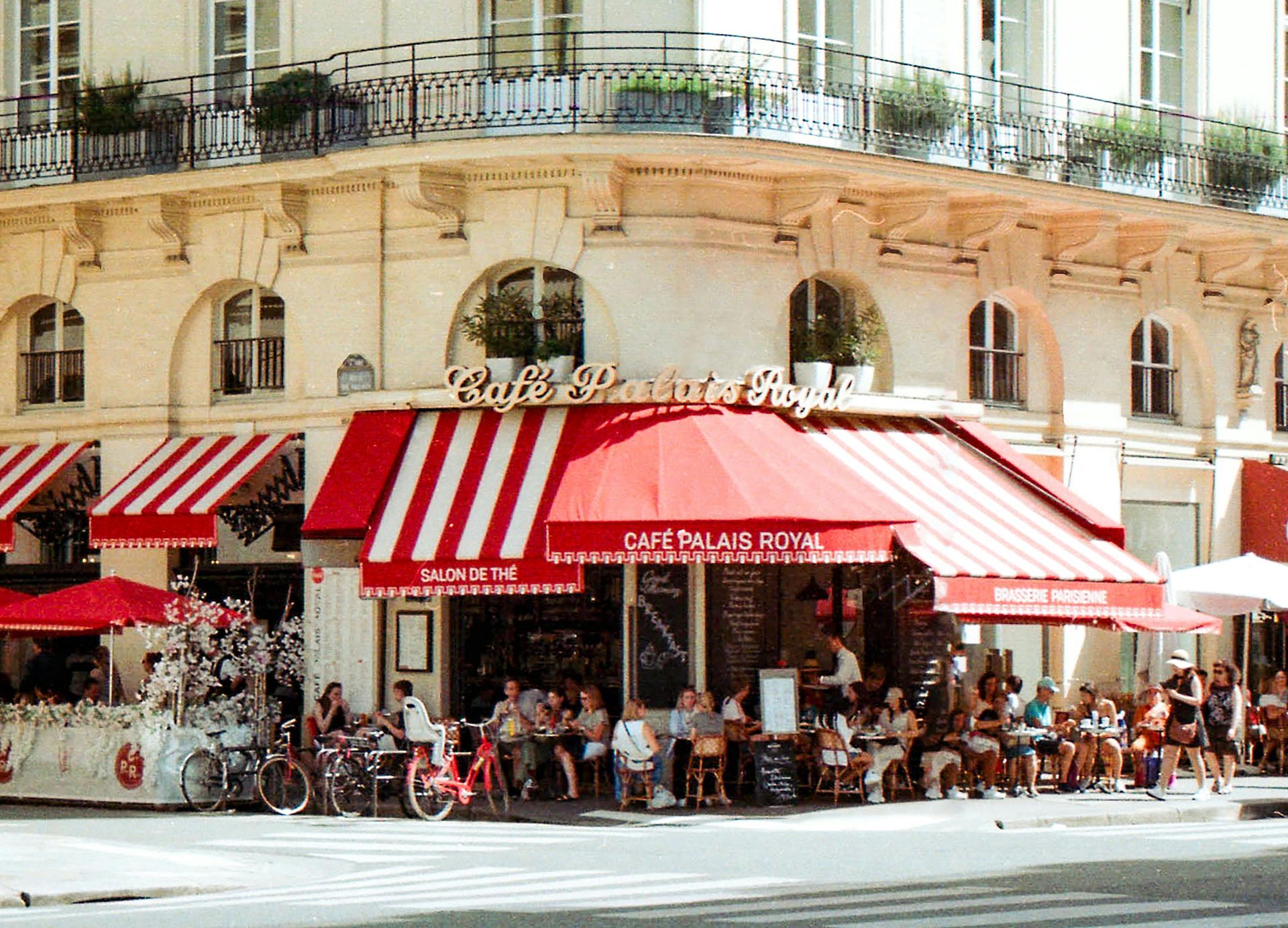 A corner café with red and white striped awnings, outdoor seating, and patrons on a sunny street.