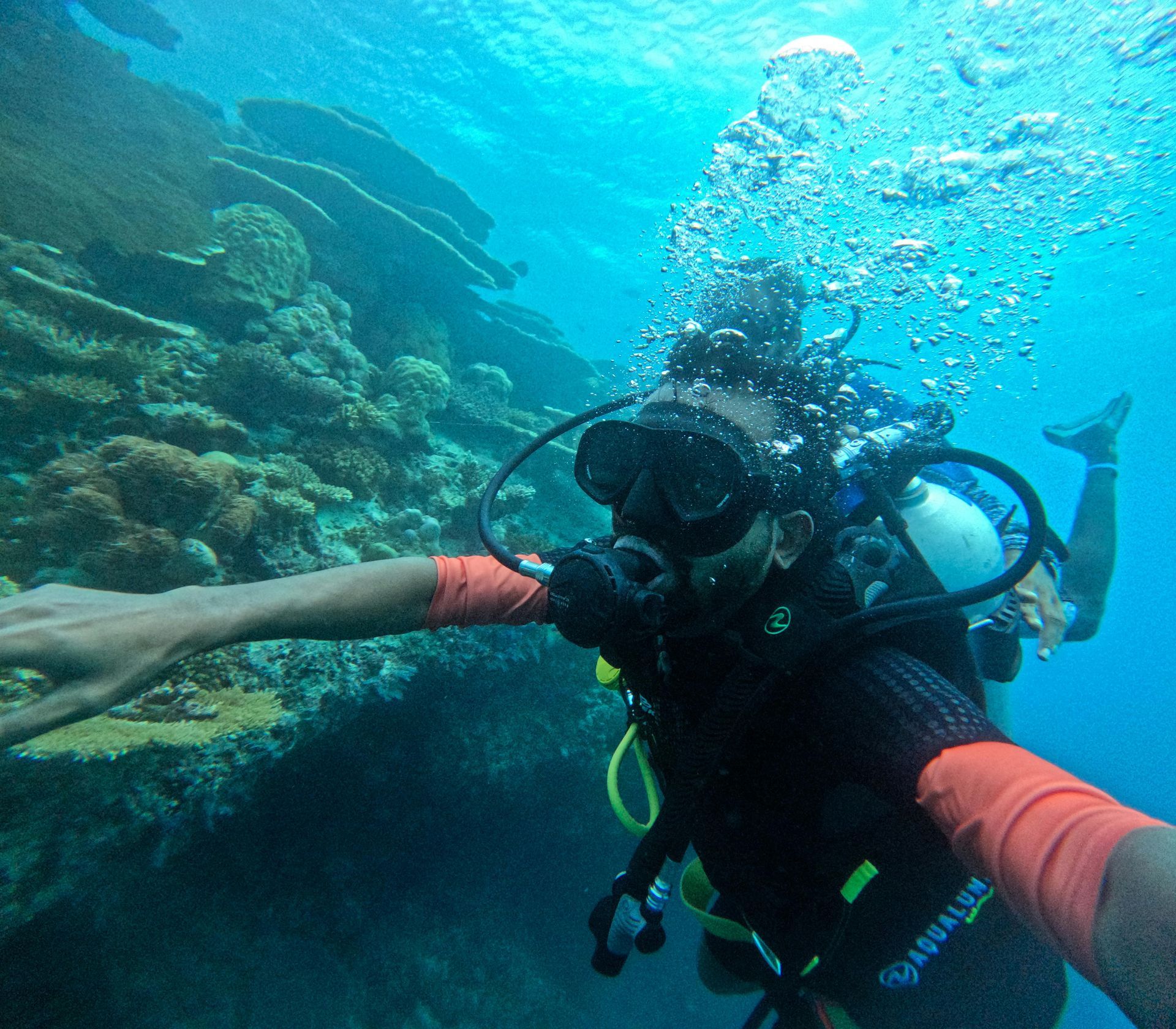 Scuba diver underwater, arms outstretched, near coral reef; bubbles rising. maldives