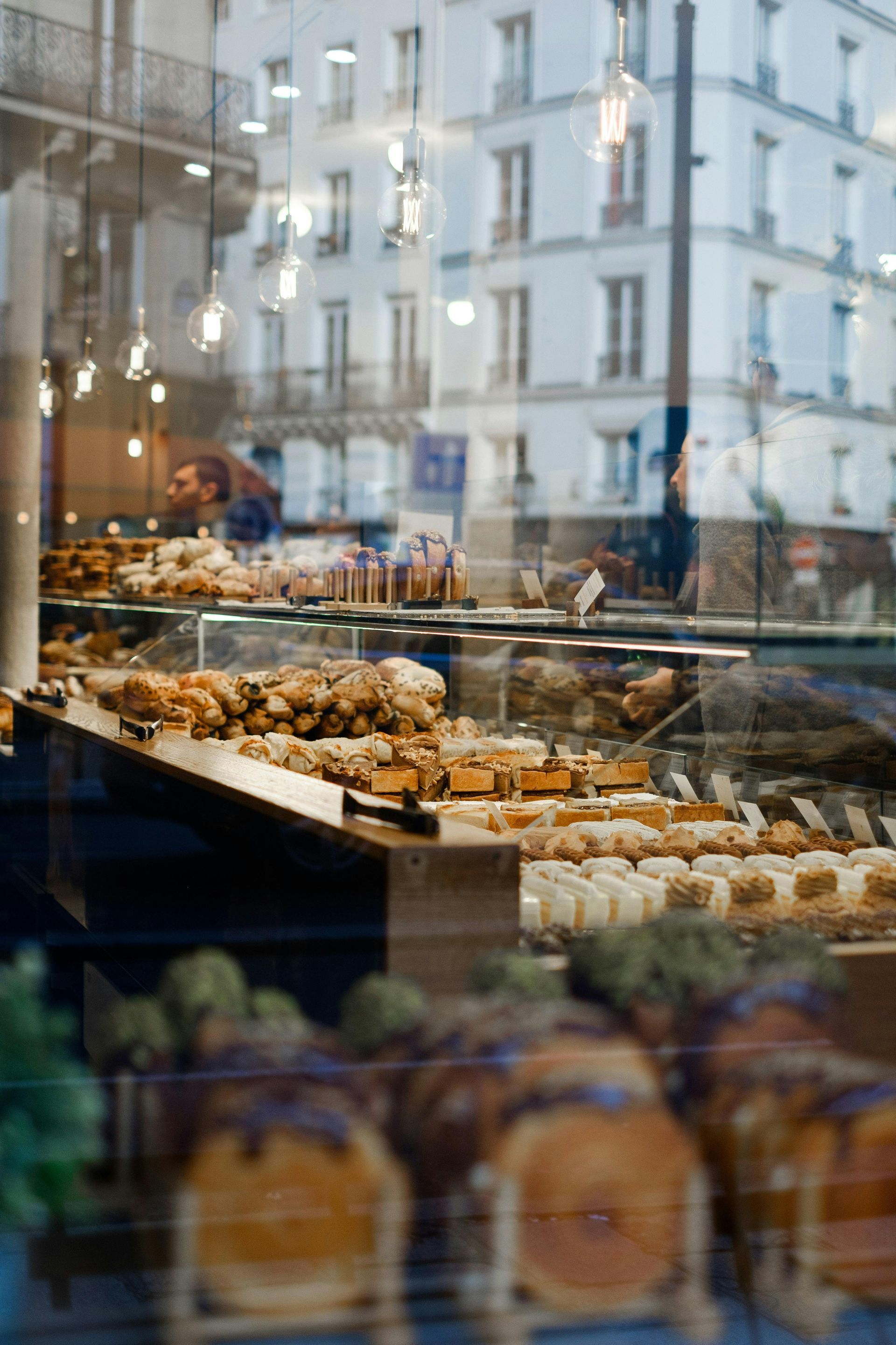 A display case inside a bakery filled with various pastries, with a view of a city street reflected in the window.