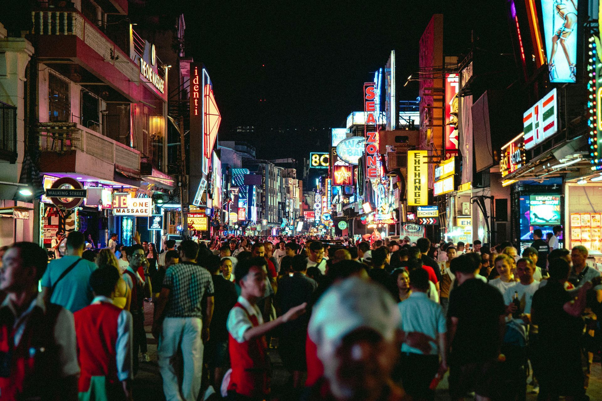 A dense crowd fills a street at night, illuminated by bright, colorful neon signs and glowing storefronts. Bangkok Thailand