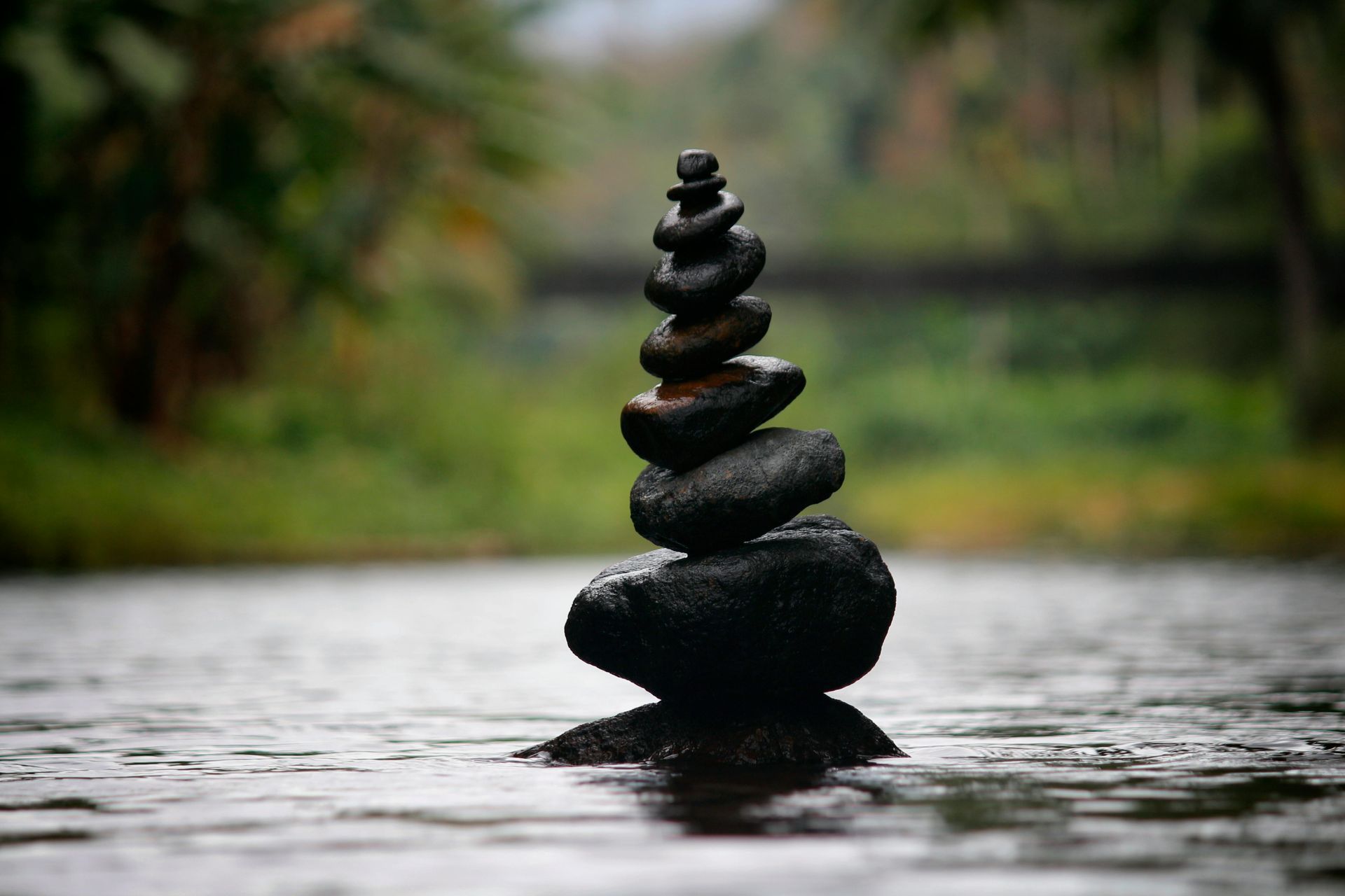 Stack of dark stones balanced in water, with blurred green forest background.