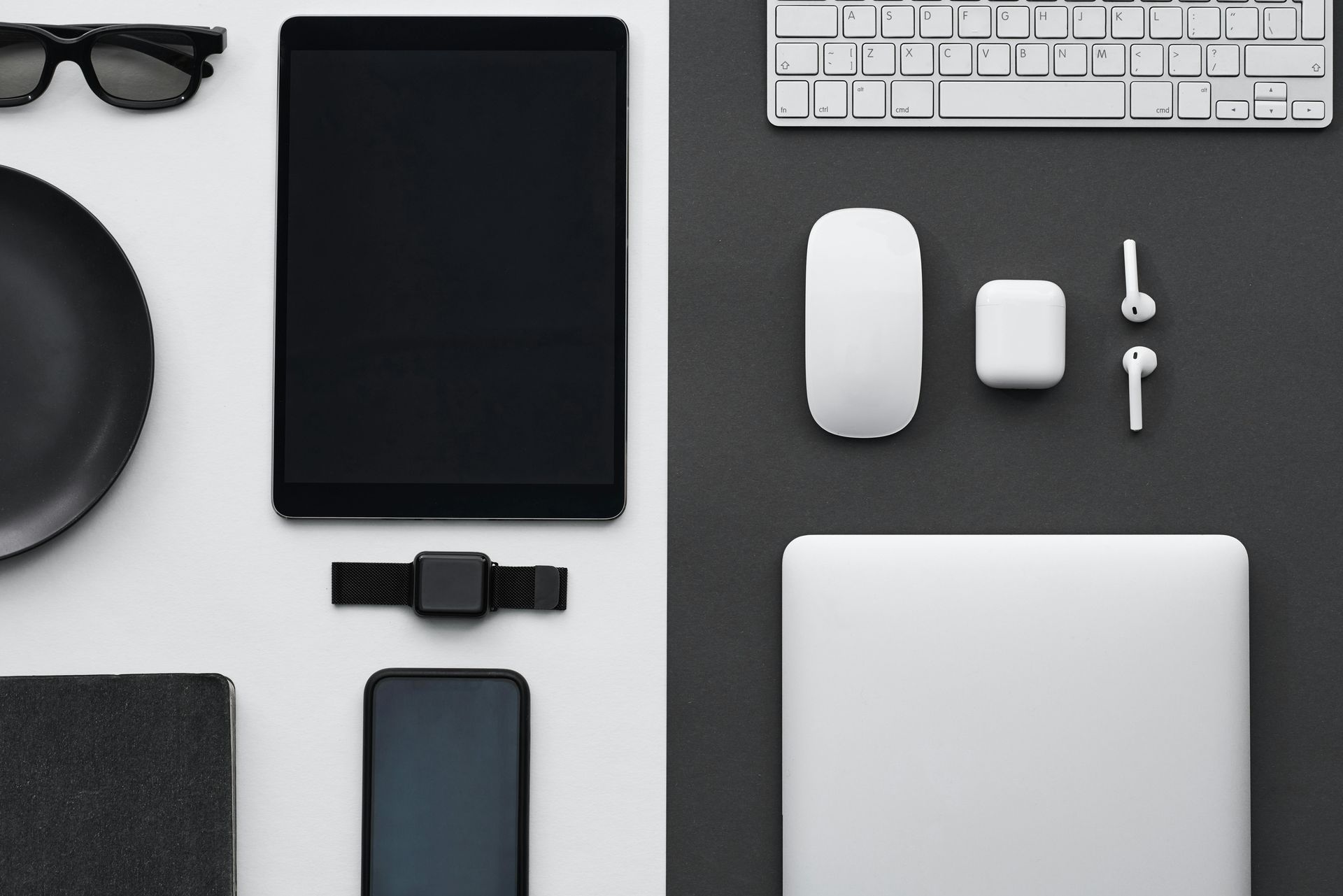 Black and white office setup with tech devices: tablet, keyboard, laptop, phone, watch, mouse, and earbuds.